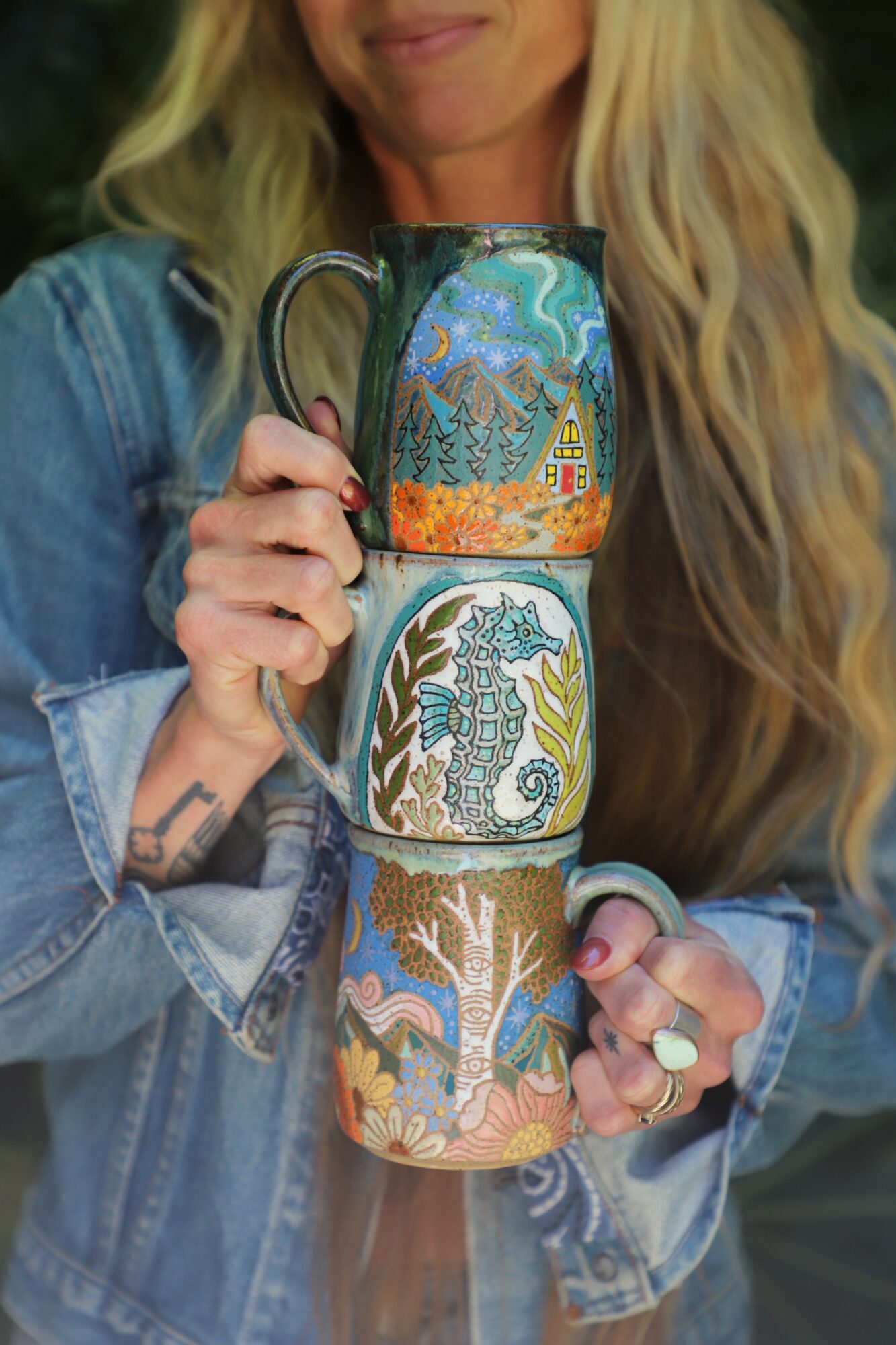 Person holding three decorated mugs, wearing a denim jacket, with long blonde hair partially visible.