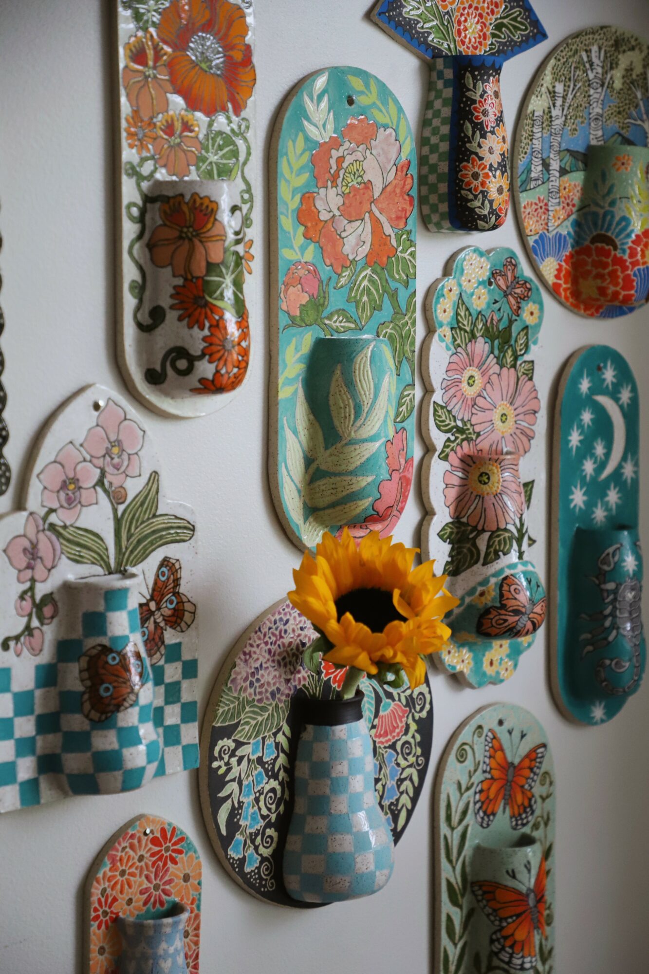 Decorative painted wooden objects with floral and geometric patterns, some with vases and a sunflower, arranged on a wall.