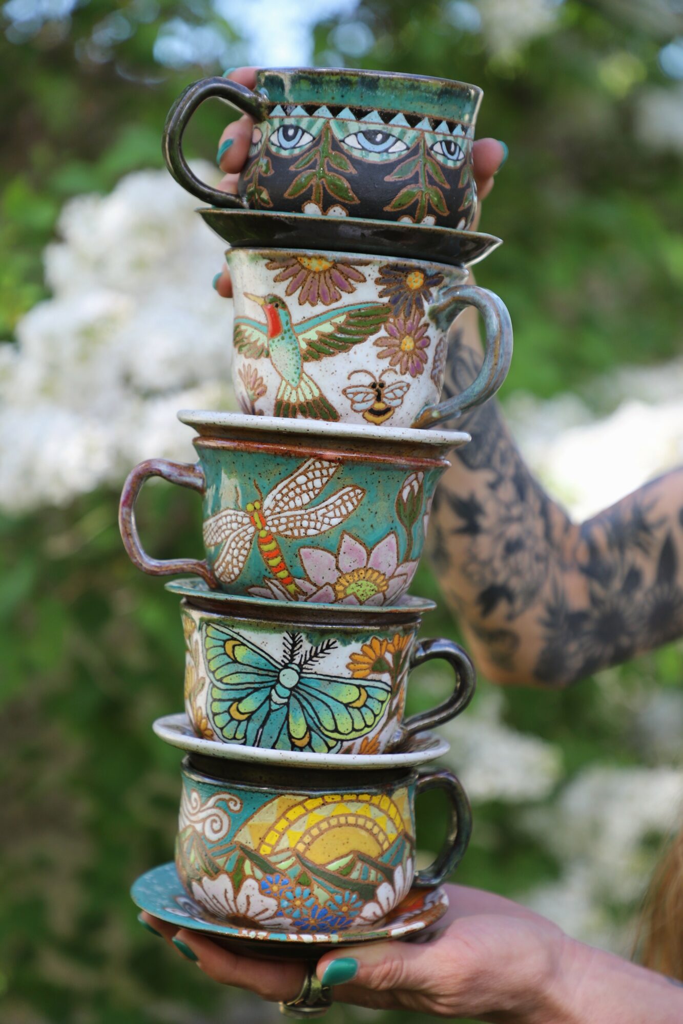 Stacked colorful cups with floral and butterfly patterns held by a person outdoors, blurred greenery in background.