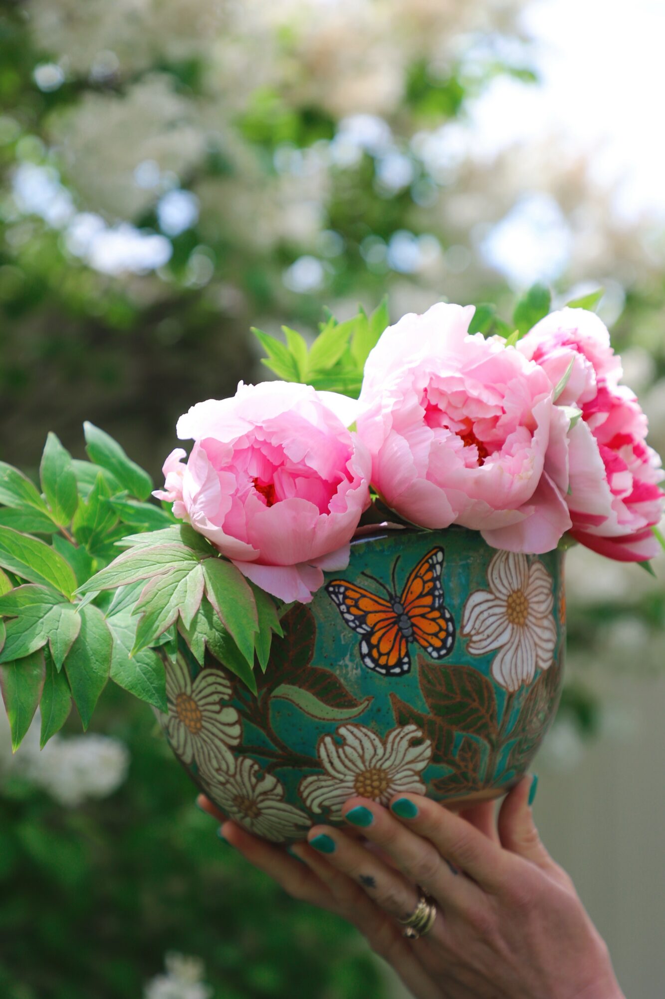 Person holding a decorative vase with pink peonies and green leaves outdoors, blurred background of trees and sky.