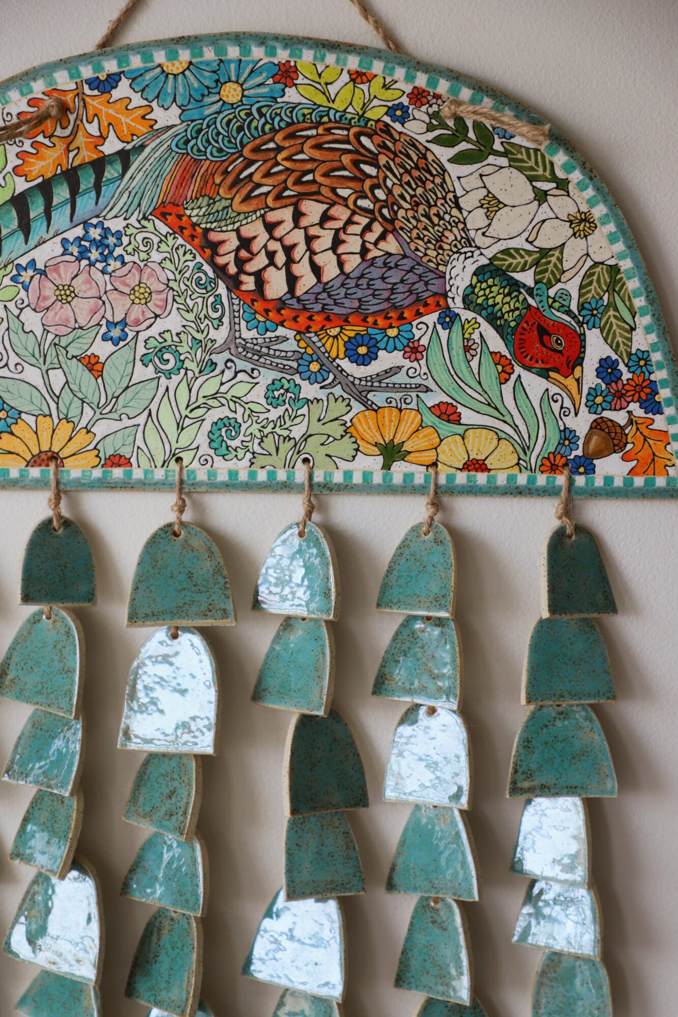 Decorative wall hanging with a colorful bird and floral embroidery, featuring hanging ceramic pieces in shades of green and white.