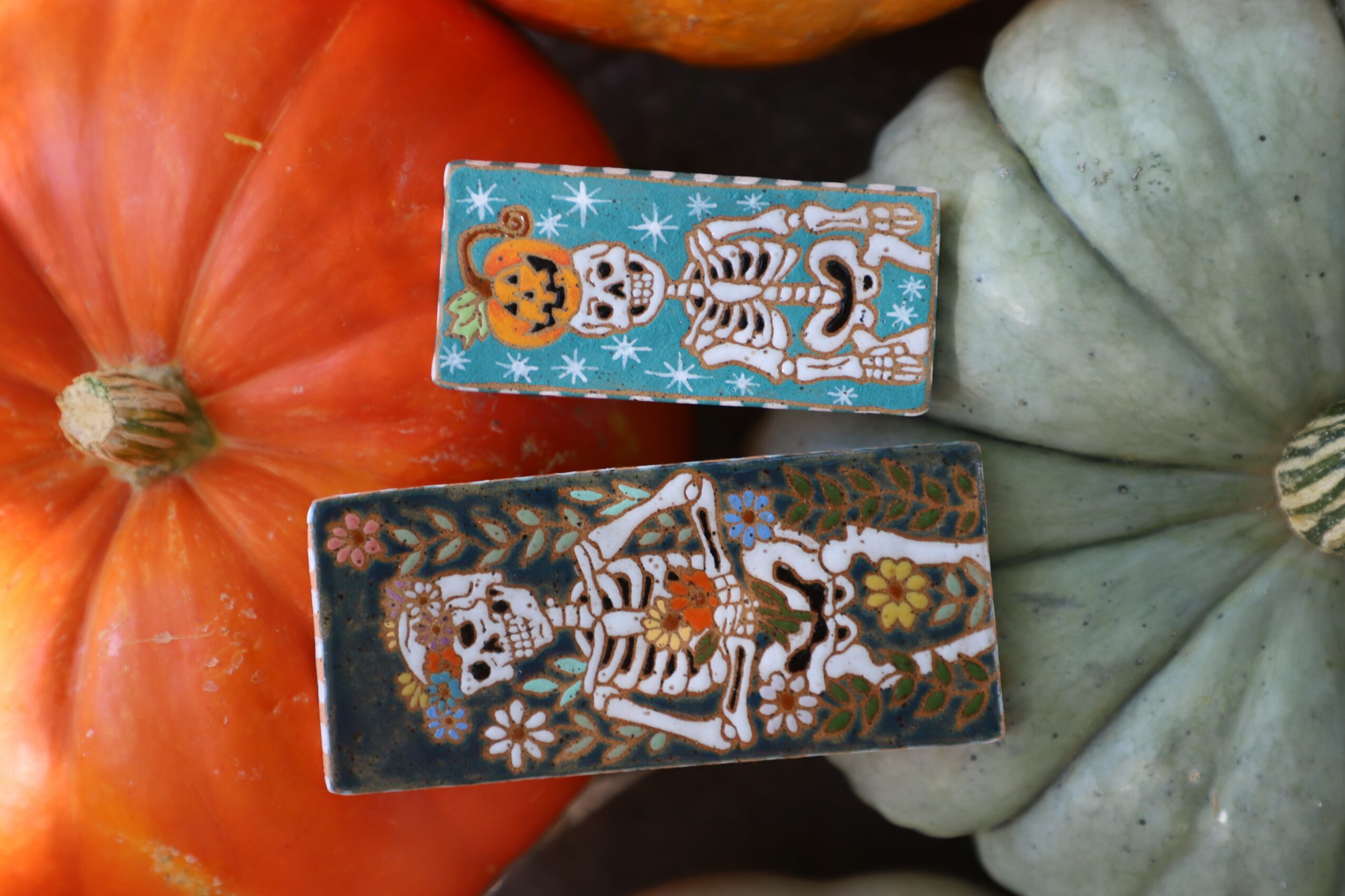 Two decorated matchboxes placed on pumpkins, one with a blue background and Halloween-themed skulls, the other with a dark background and skeletons.