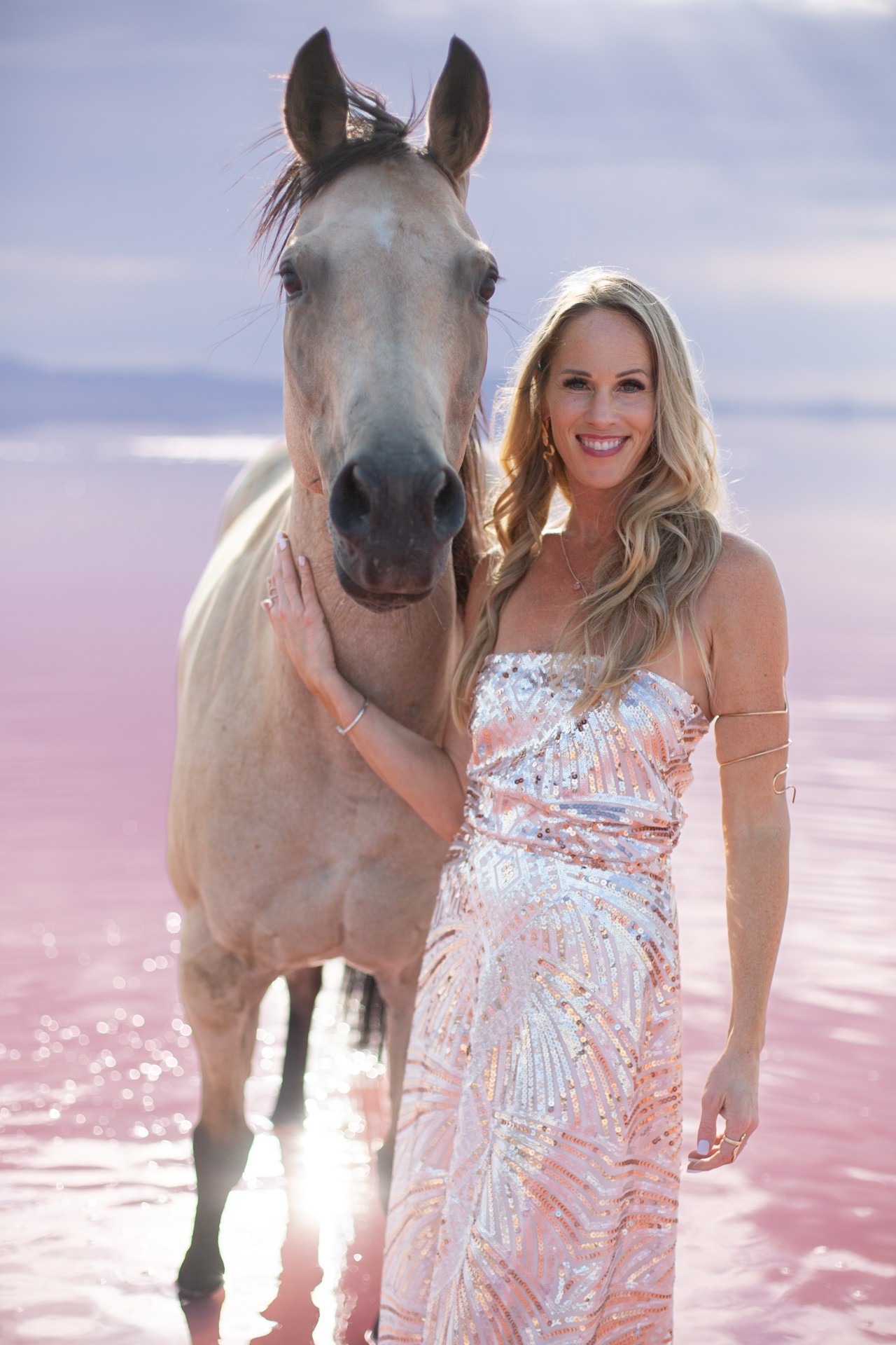 Woman with long blonde hair smiling next to a light-colored horse on a pink beach, ocean in background