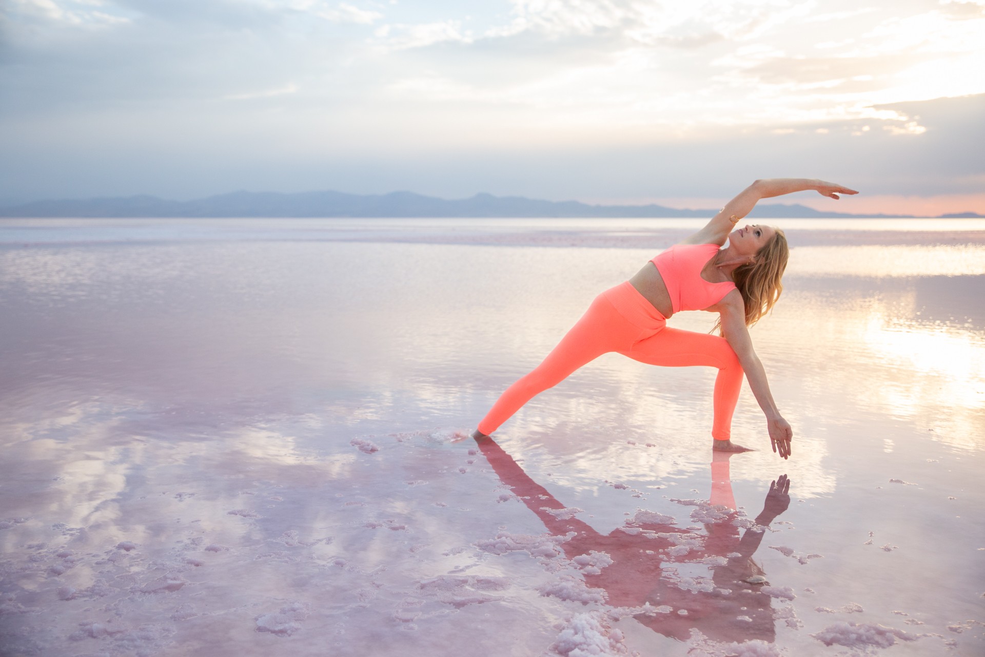 Person in pink workout clothes performing a yoga pose on a reflective surface with a cloudy sky and distant mountains.