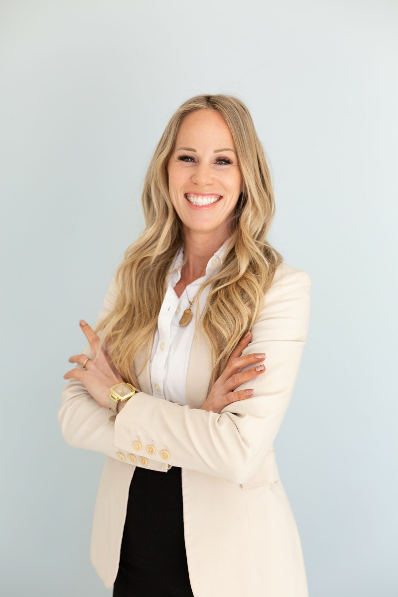 Smiling woman with long blonde hair, wearing a white blazer and black pants, standing with arms crossed against a light background.
