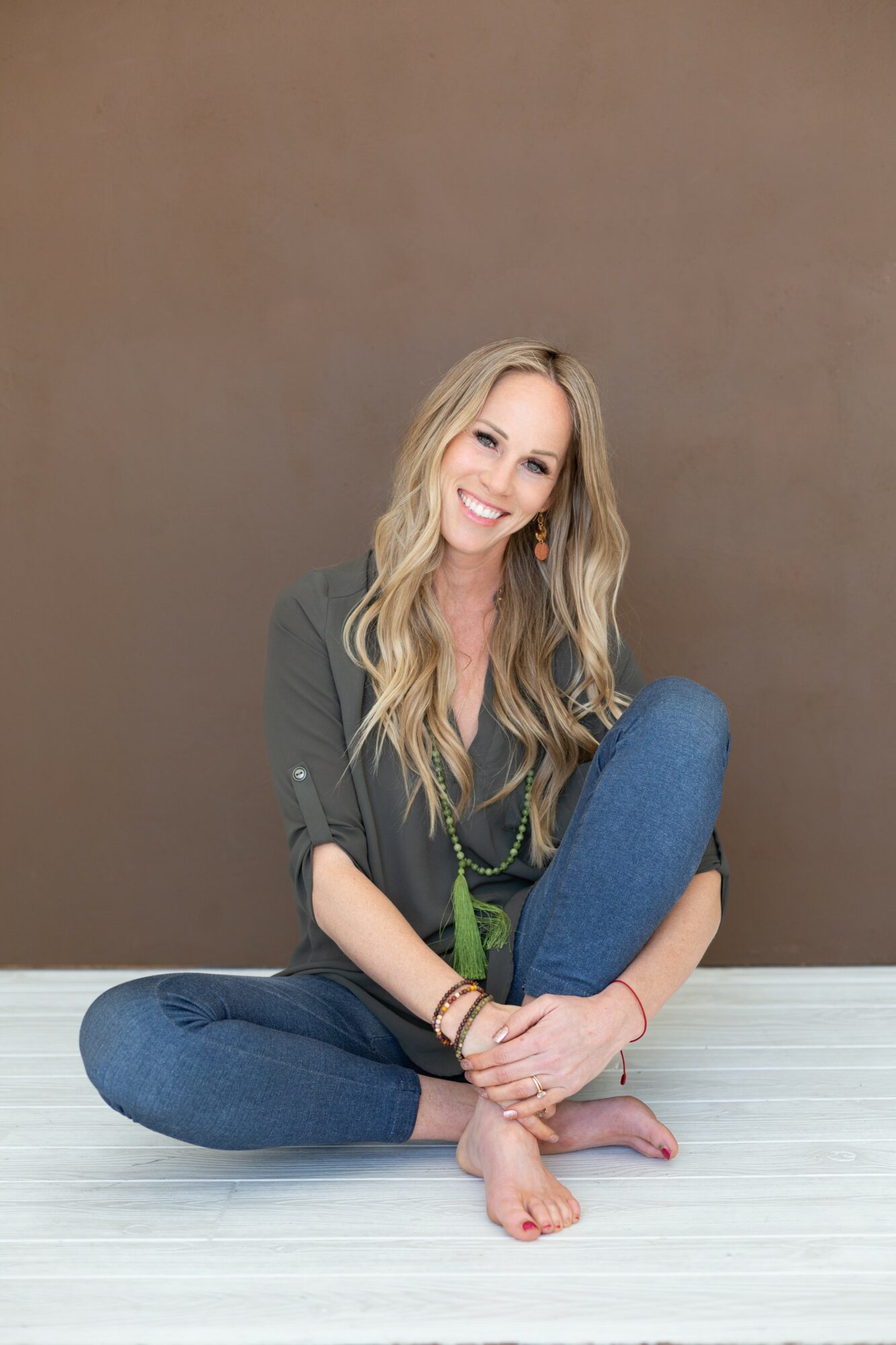 Woman with long blonde hair sitting cross-legged on floor, smiling, against a plain brown wall.