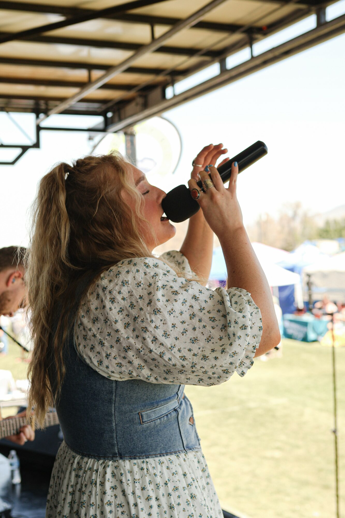 Woman singing into a microphone outdoors under a roof, with a grassy area and tents in the background.