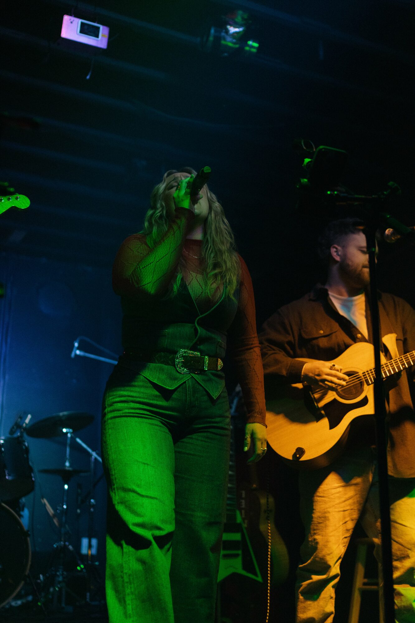 Two musicians perform on stage, one singing and the other playing guitar, with stage lighting and musical equipment visible.