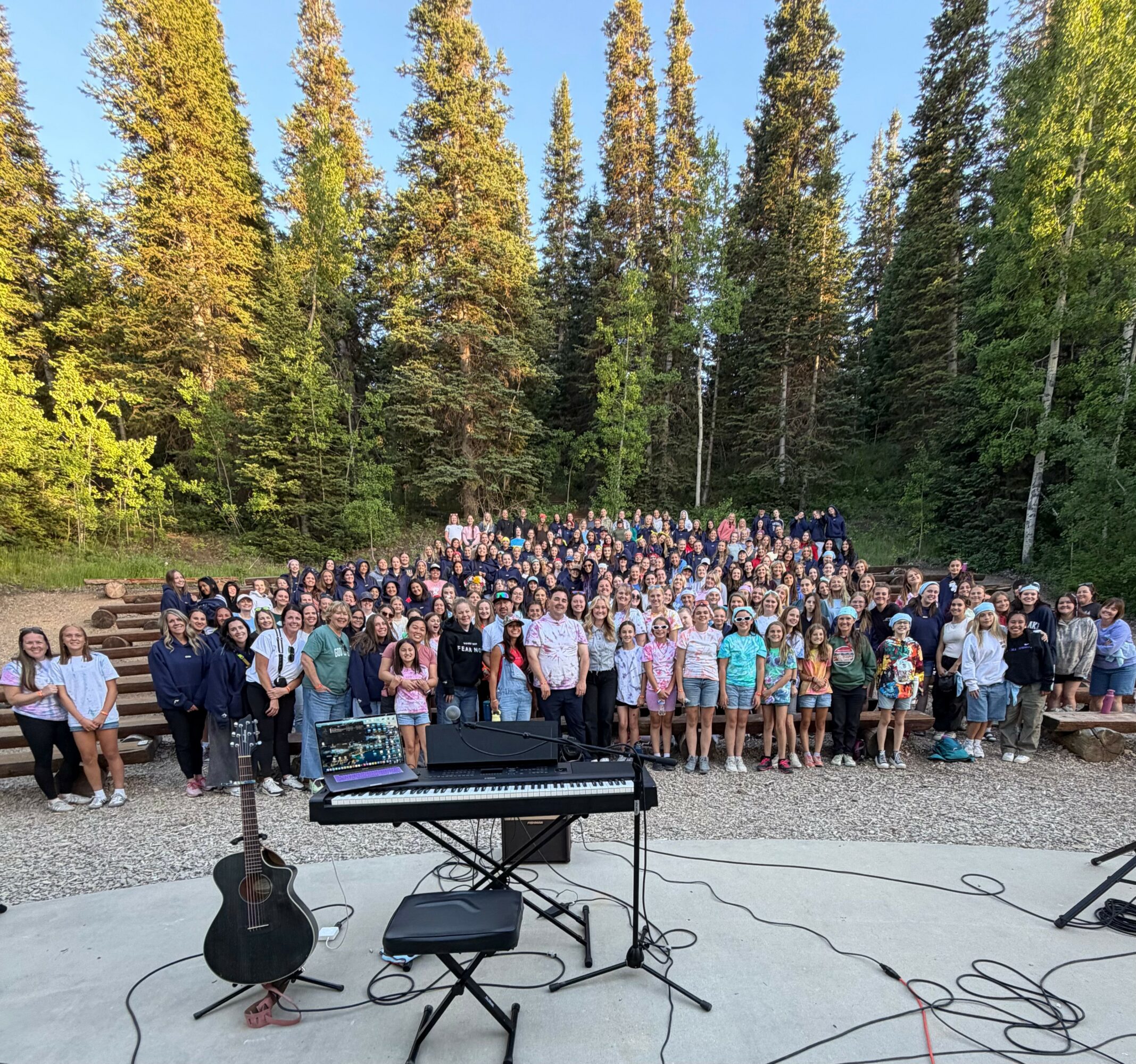 Large group of people gathered outdoors in front of tall trees, with a piano and guitar on stage, under a clear sky.