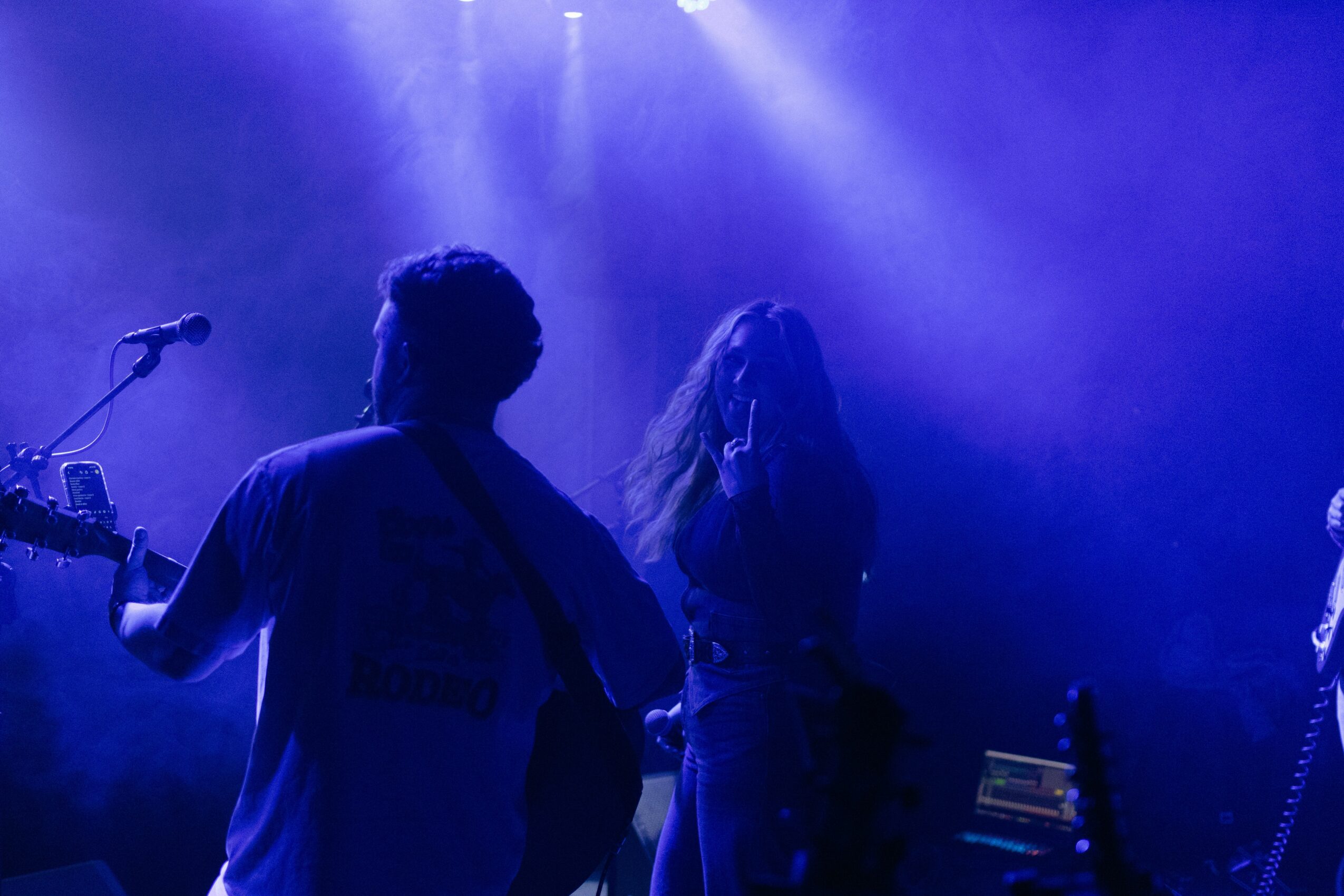 Silhouettes of two people on stage with blue lighting, one holding a guitar and the other covering their face.