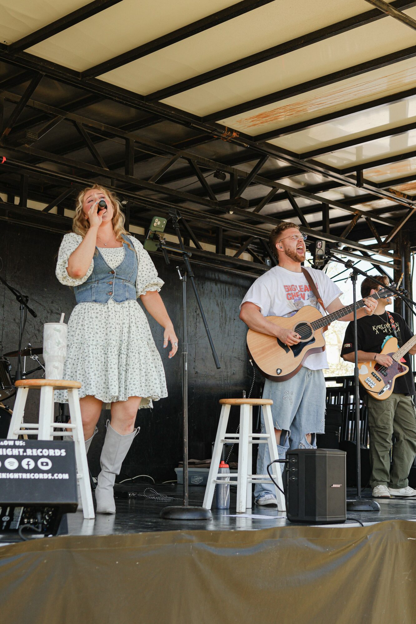 Three musicians perform on stage with guitars and microphones, under a metal roof structure, with a stool and equipment nearby.