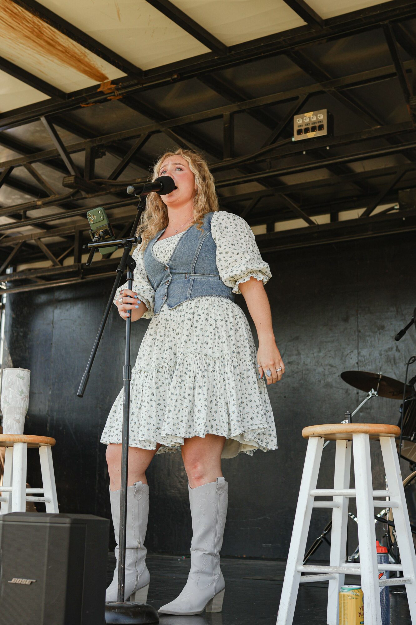 Woman singing into a microphone on stage, wearing a white dress with puffy sleeves and high boots, with stools nearby.