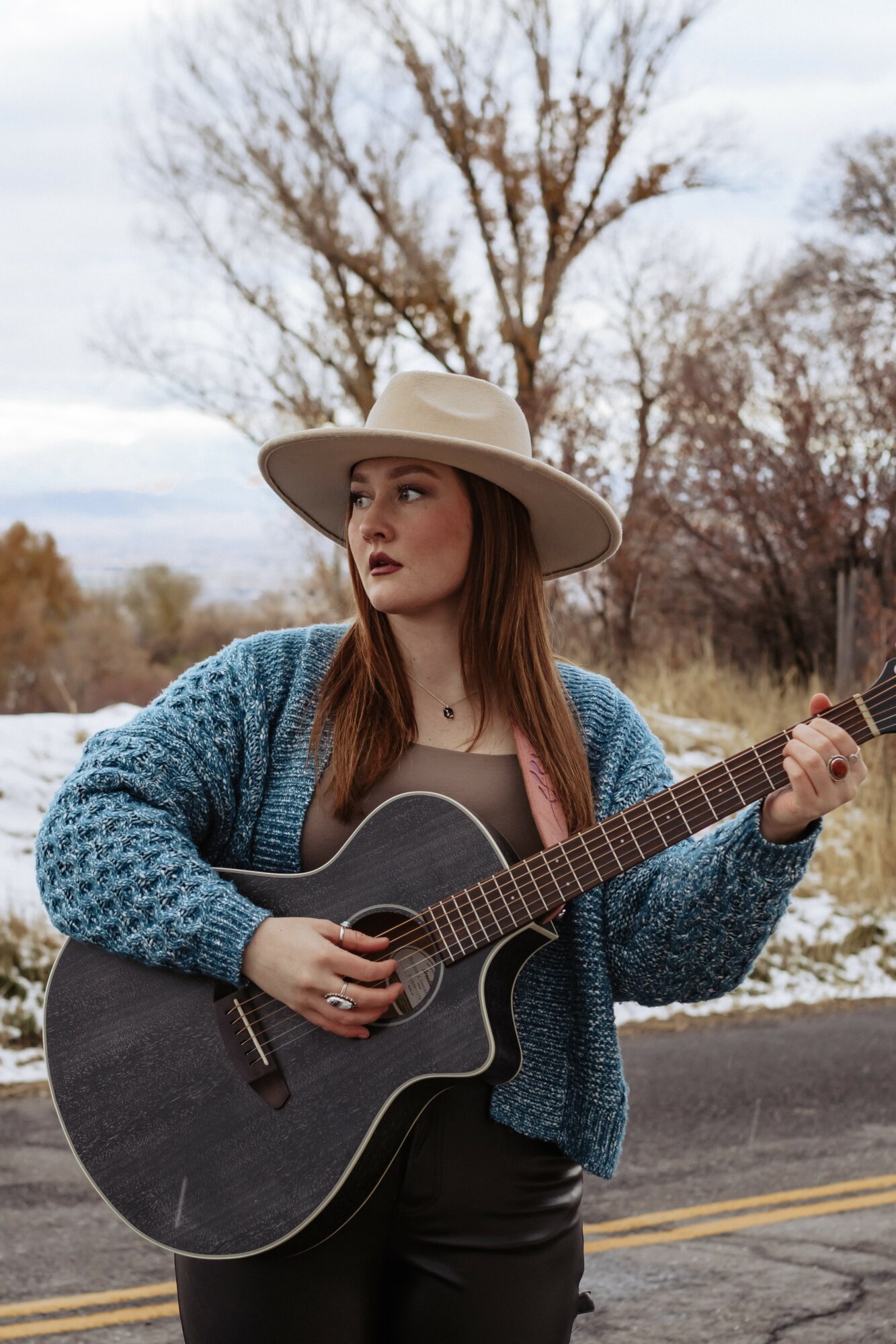 Young woman with long hair wearing a wide-brimmed hat and blue sweater, holding a black acoustic guitar outdoors.