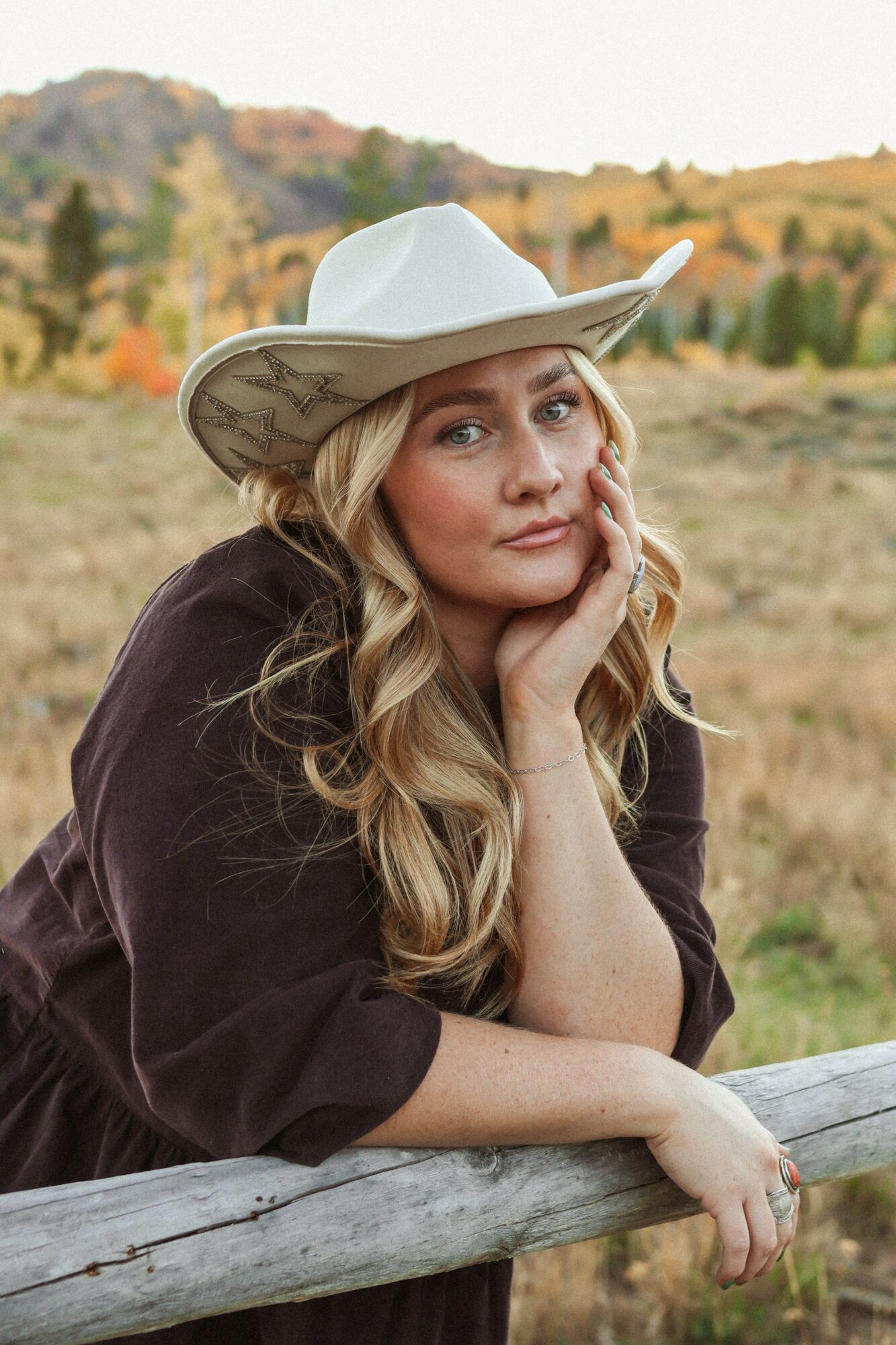 Woman with long blonde hair wearing a white cowboy hat, resting her chin on her hand outdoors in a rural landscape.