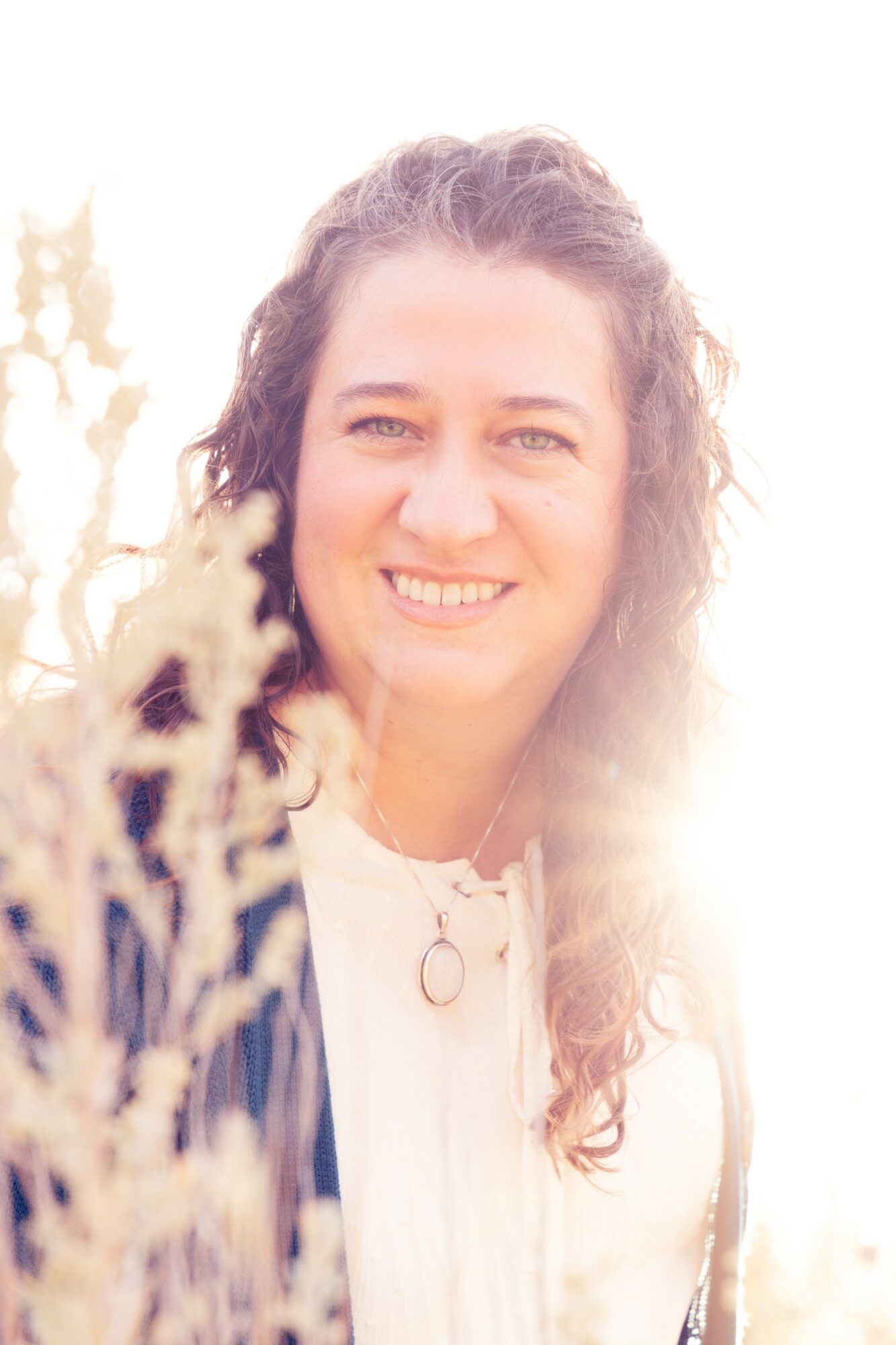 Young woman with curly hair smiling outdoors, sunlight behind her, wearing a white top with a necklace.