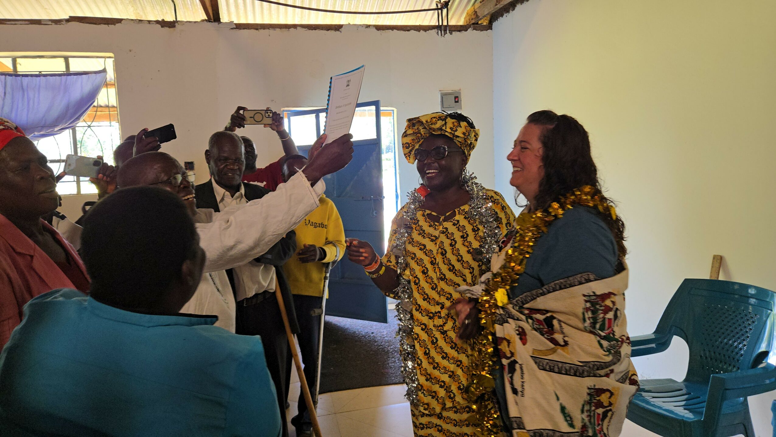 Group of people indoors, some exchanging documents, with a woman in traditional attire and a woman with a shawl, in a room with chairs and windows.
