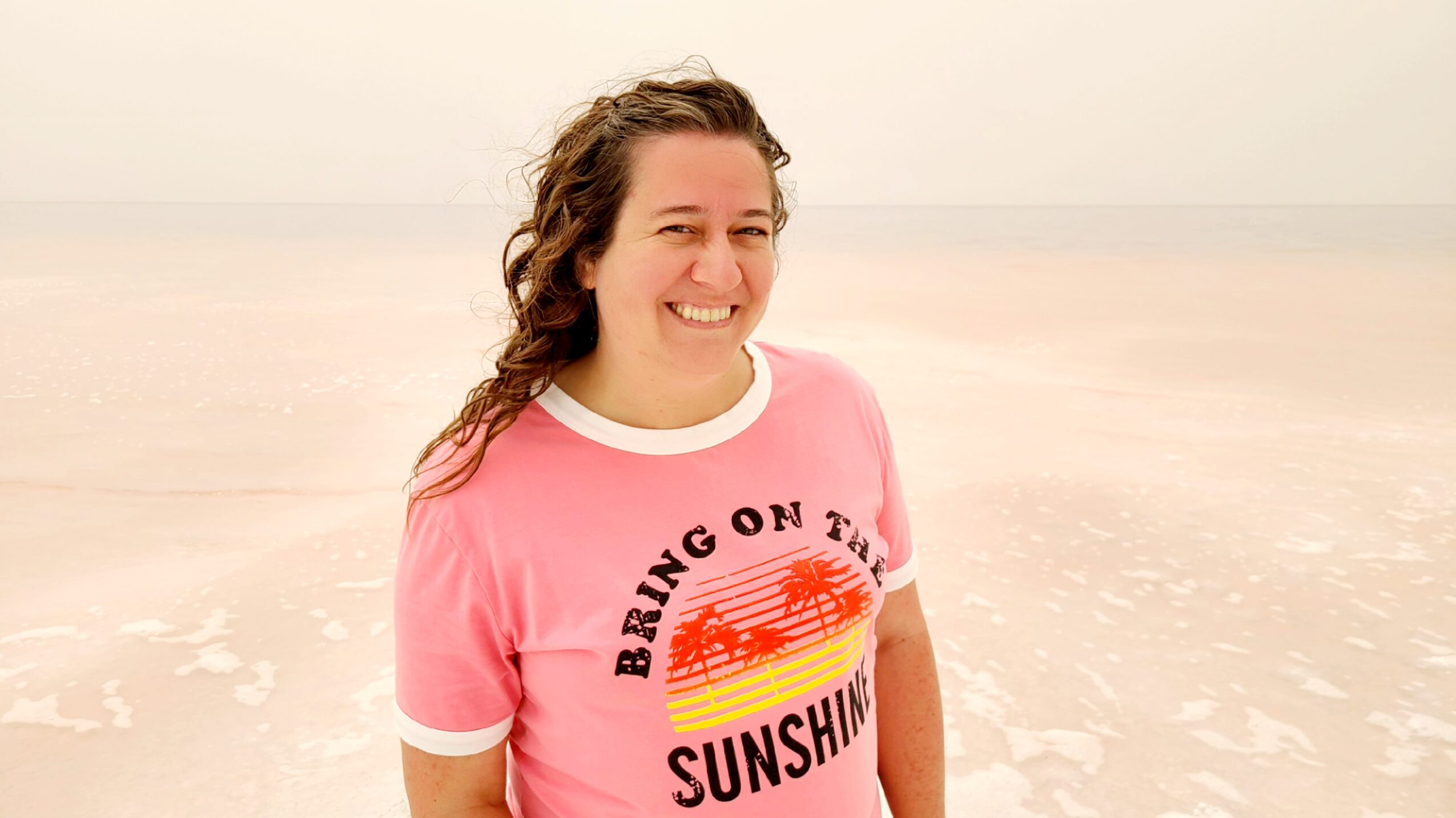 Smiling woman with curly hair wearing a pink T-shirt on a beach, ocean in background.