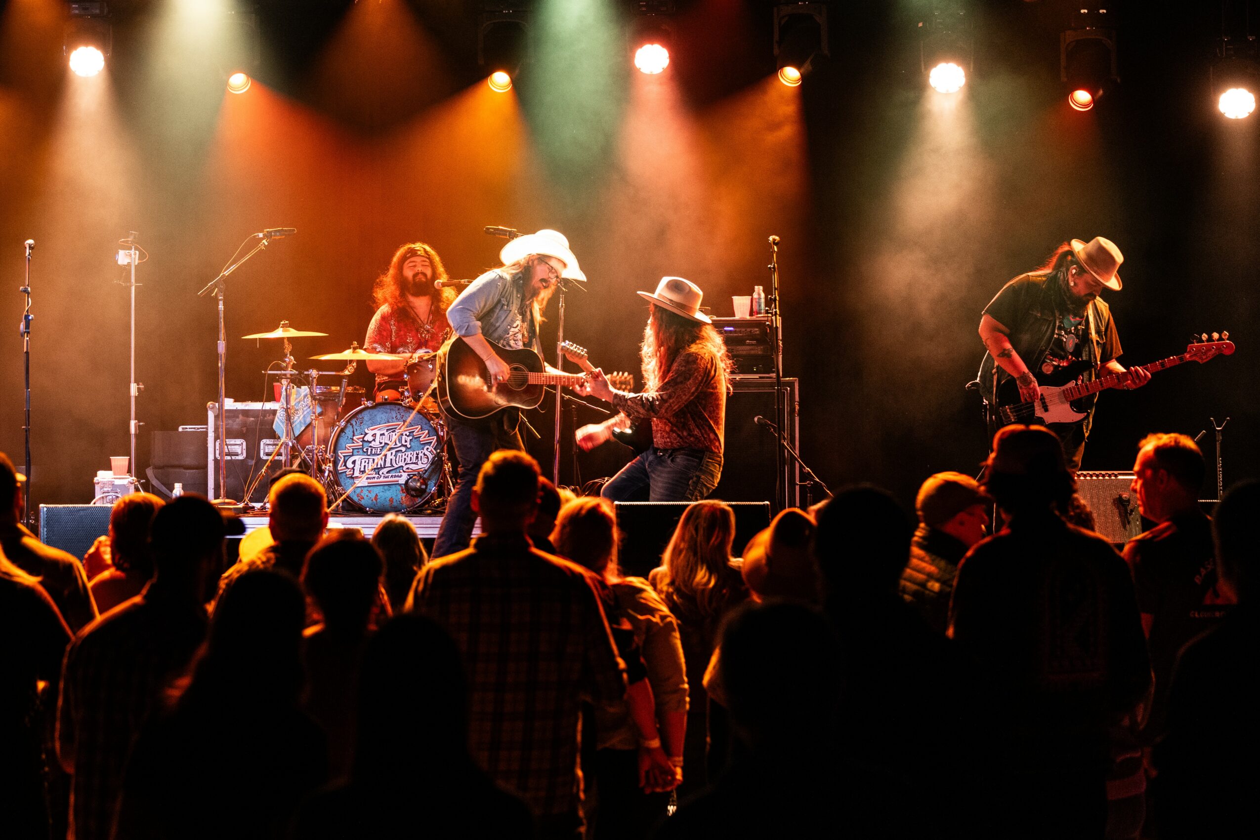 Band performs on stage with audience in foreground, colorful stage lights overhead, musicians playing instruments, some wearing hats.