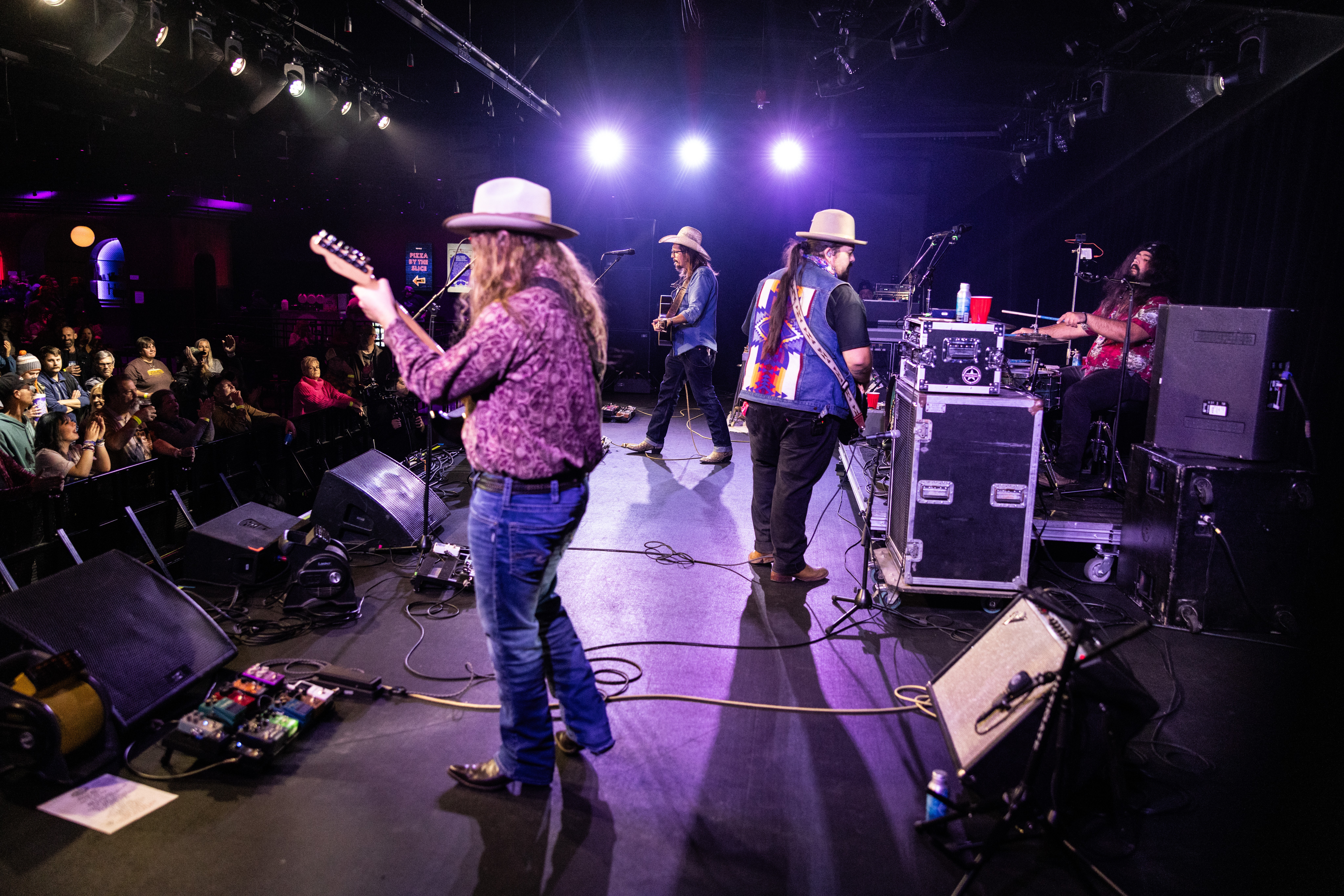 Band performs on stage with musicians playing guitars and drums, audience visible in the background, stage lighting overhead.