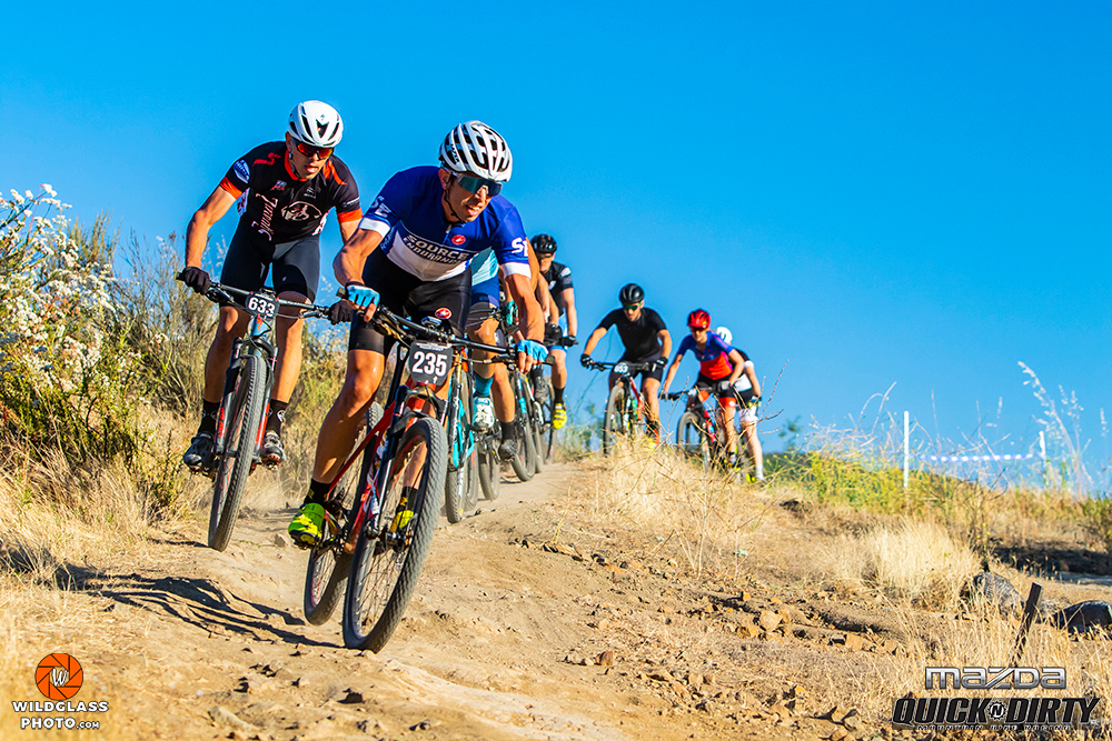 Group of cyclists riding on a dirt trail in a sunny outdoor setting with clear blue sky.