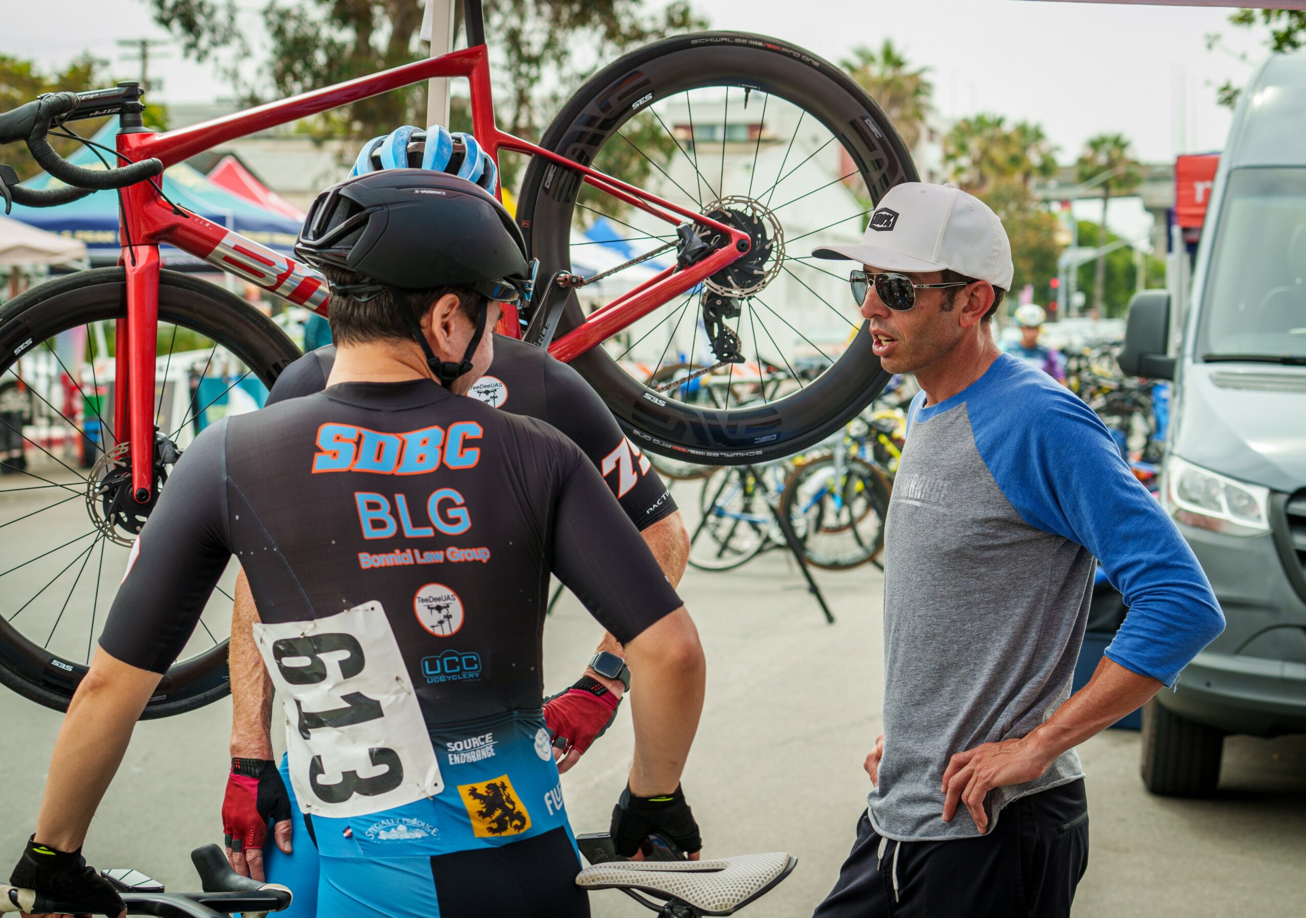 Two men talking near a red bicycle mounted on a bike rack, with other bikes and vehicles in the background.