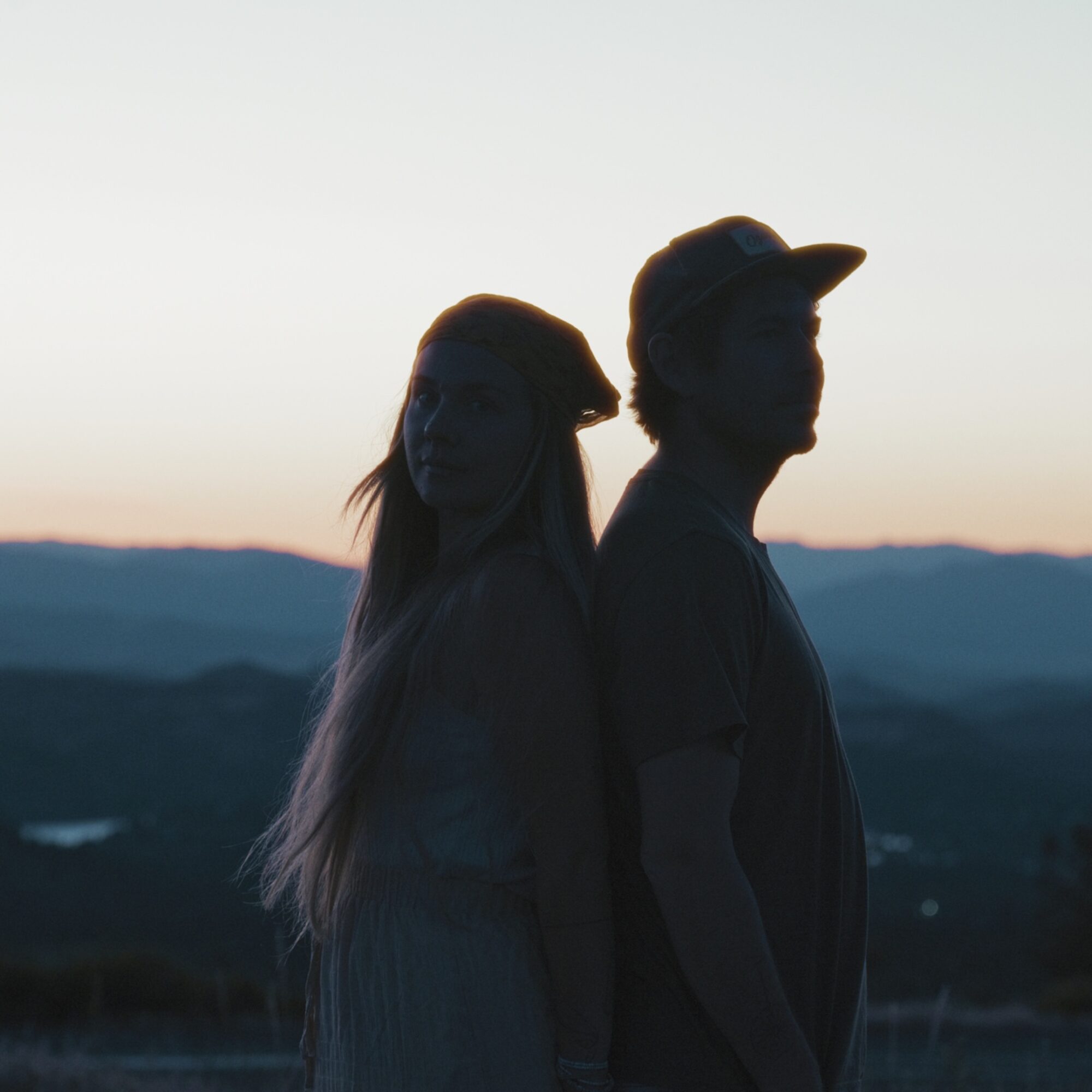 Silhouettes of a woman and a man standing outdoors during sunset, with mountains in the background.