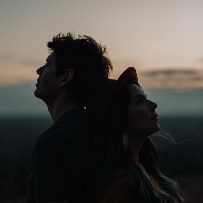 Two people stand back-to-back outdoors during sunset, with a blurred landscape background, one wearing a hat, both in profile.