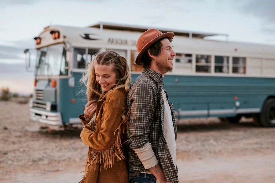 A smiling young woman and man stand outdoors near a vintage bus, with a cloudy sky in the background.
