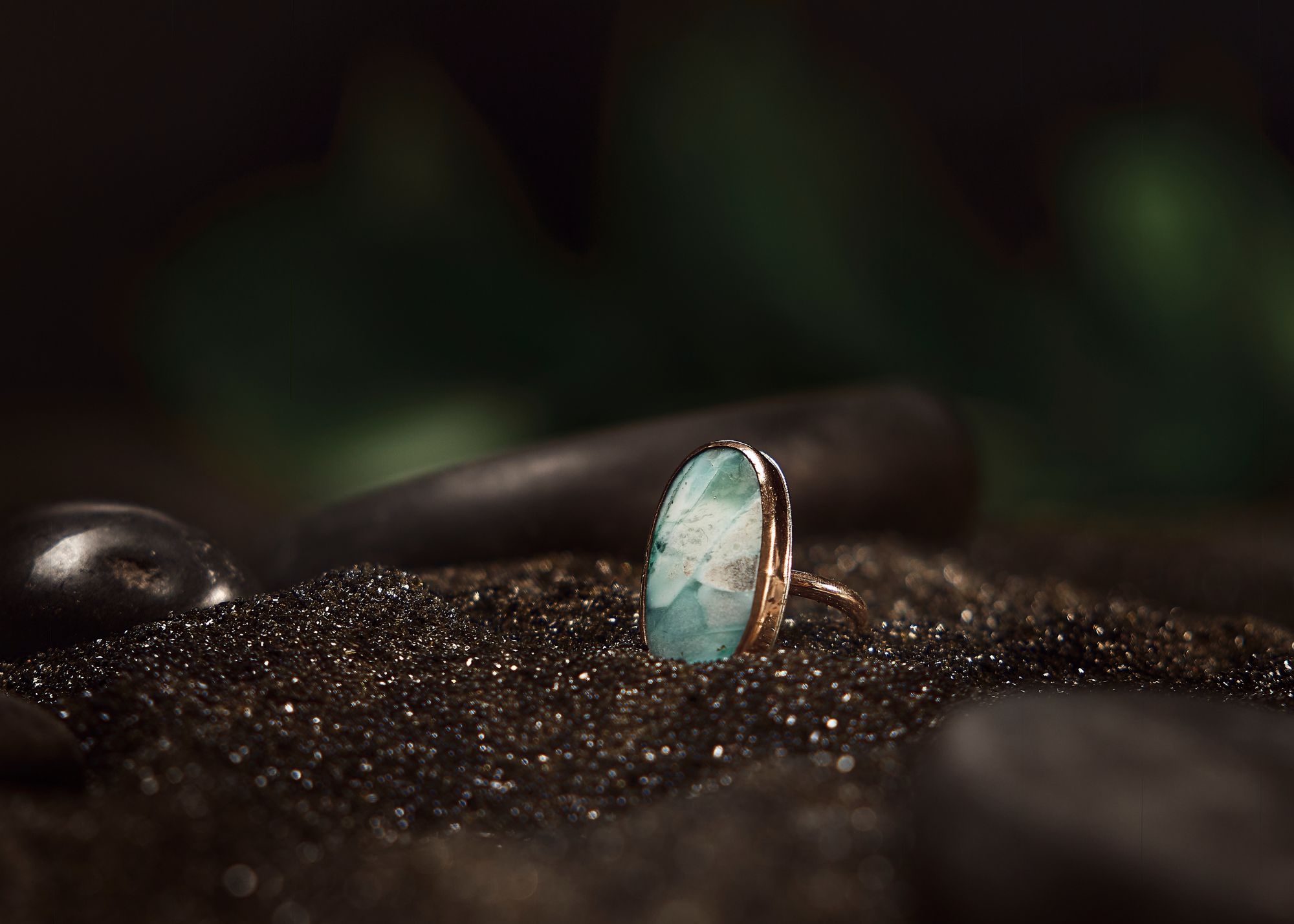 Ring with a blue and white stone on dark, textured surface with blurred background leaves.