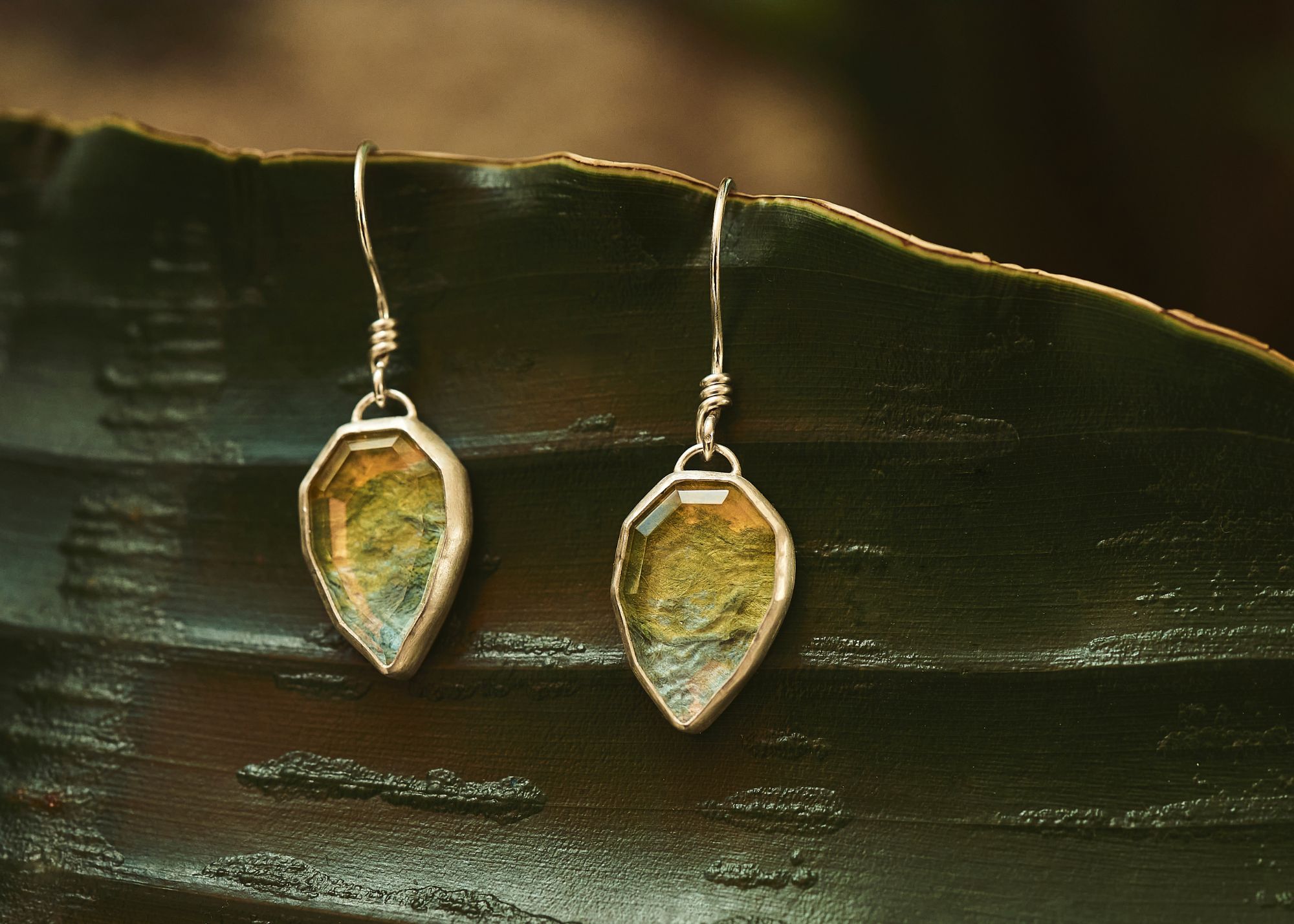 Pair of earrings with greenish stones hanging from silver hooks, resting on a dark green leaf surface.