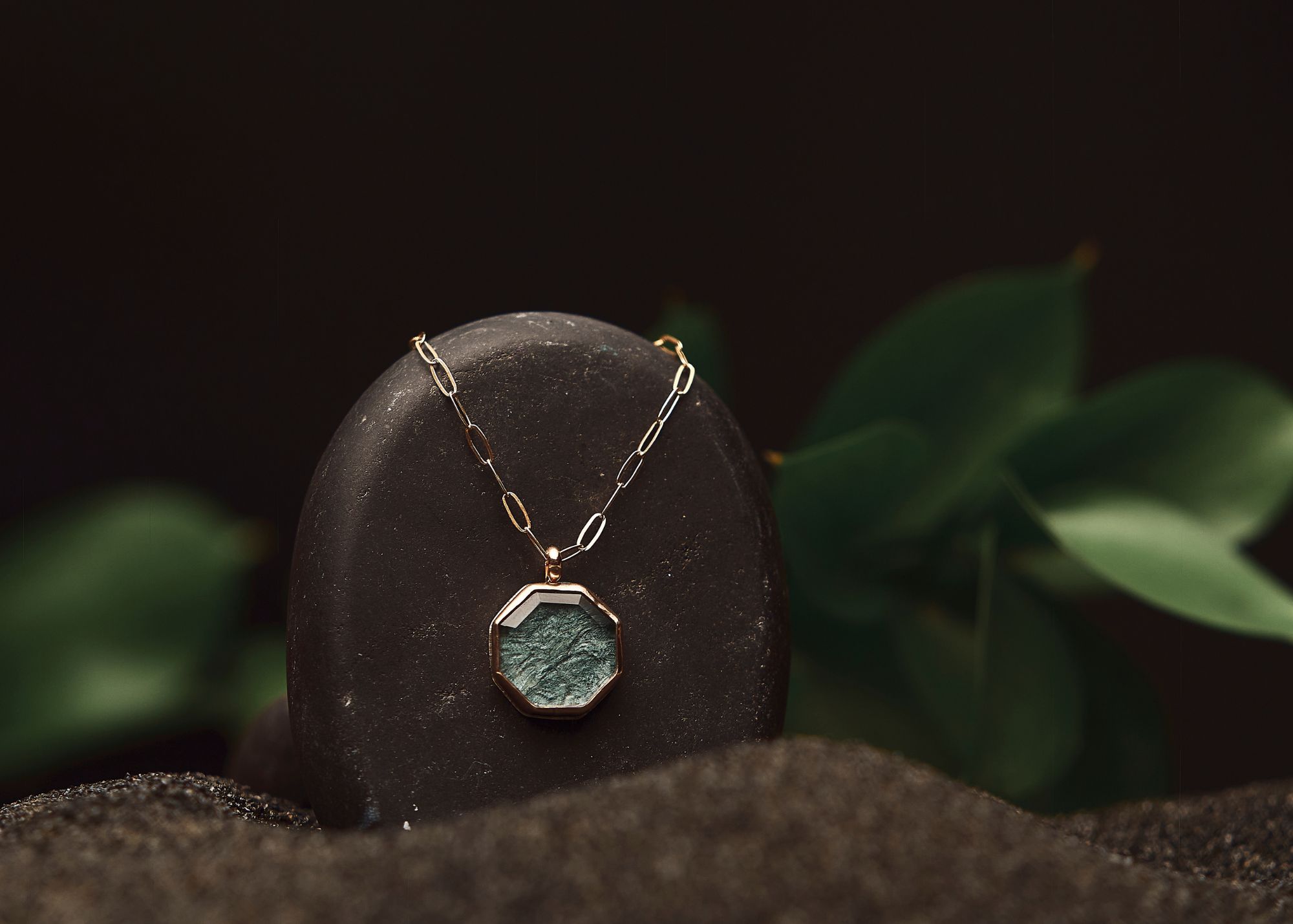 Necklace with a hexagonal pendant displayed on a black oval stand, with green leaves in the background.