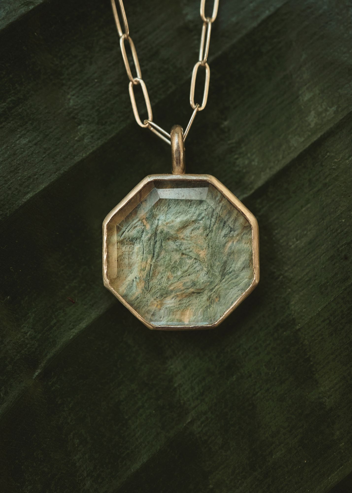 Pendant with a greenish, textured surface in an octagonal frame hanging from a chain, against a dark background.