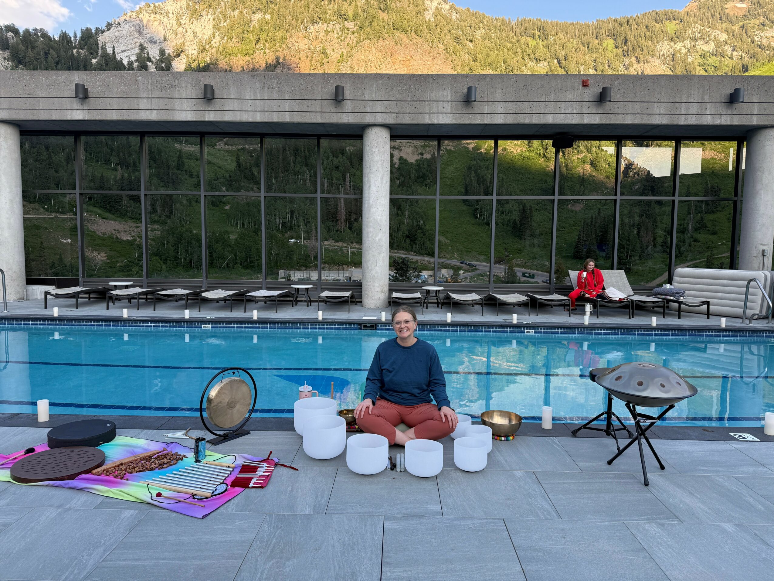Person sitting cross-legged by a pool with musical instruments and colorful mats, outdoor setting with glass windows and mountains.