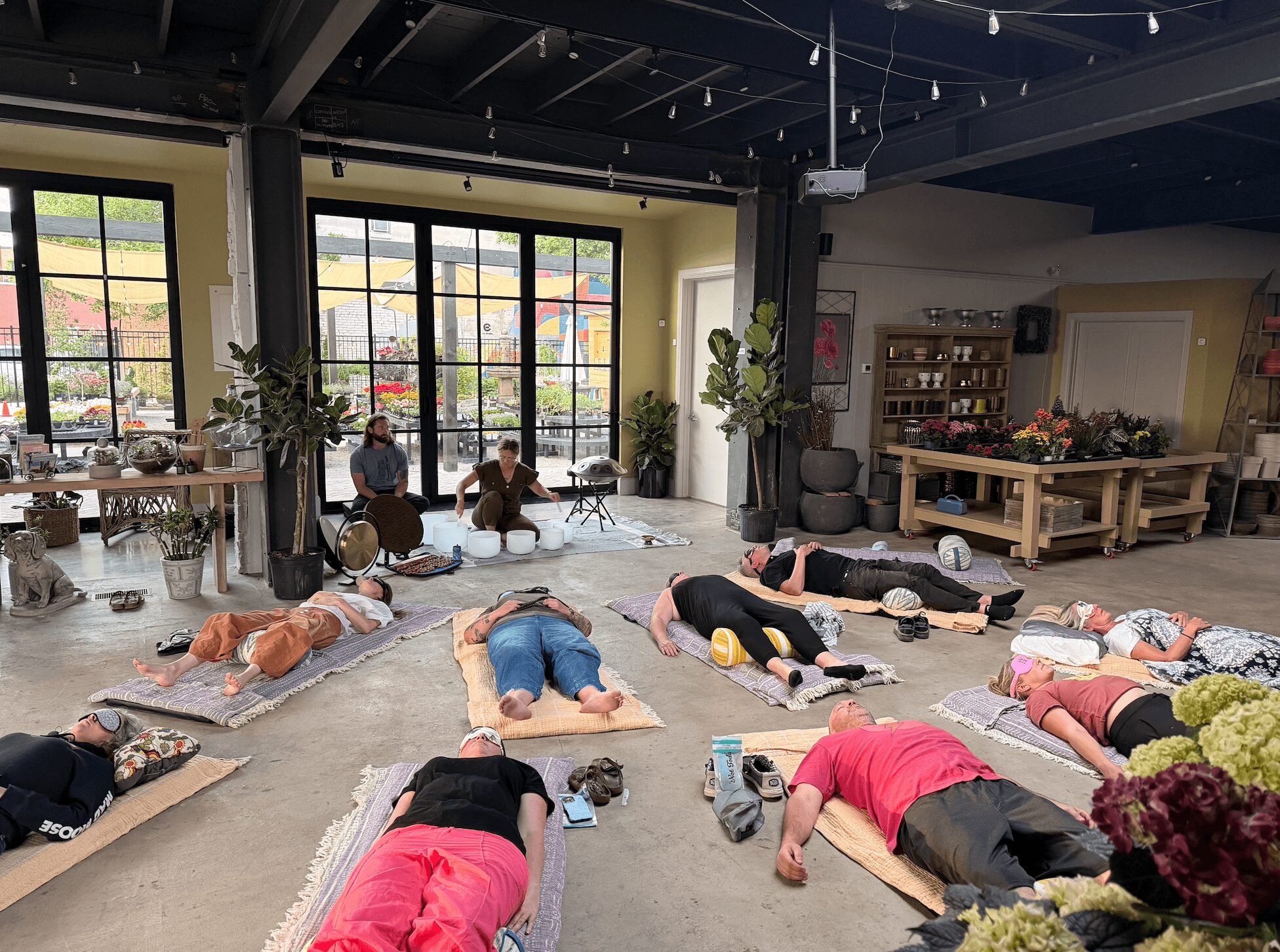 People lying on mats in a spacious room with large windows, plants, and furniture, participating in a group activity or class.