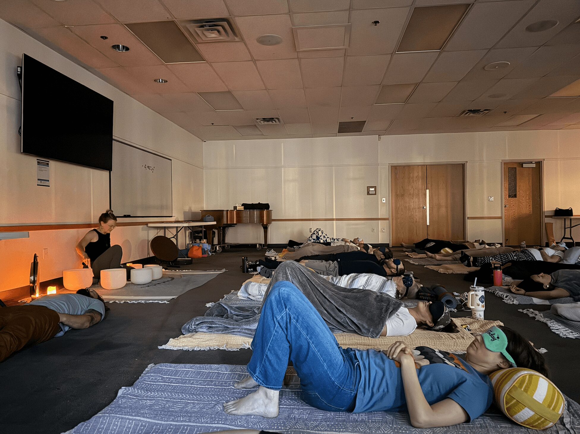 People lying on mats in a room, some resting, with a large screen on the wall, and doors in the background.