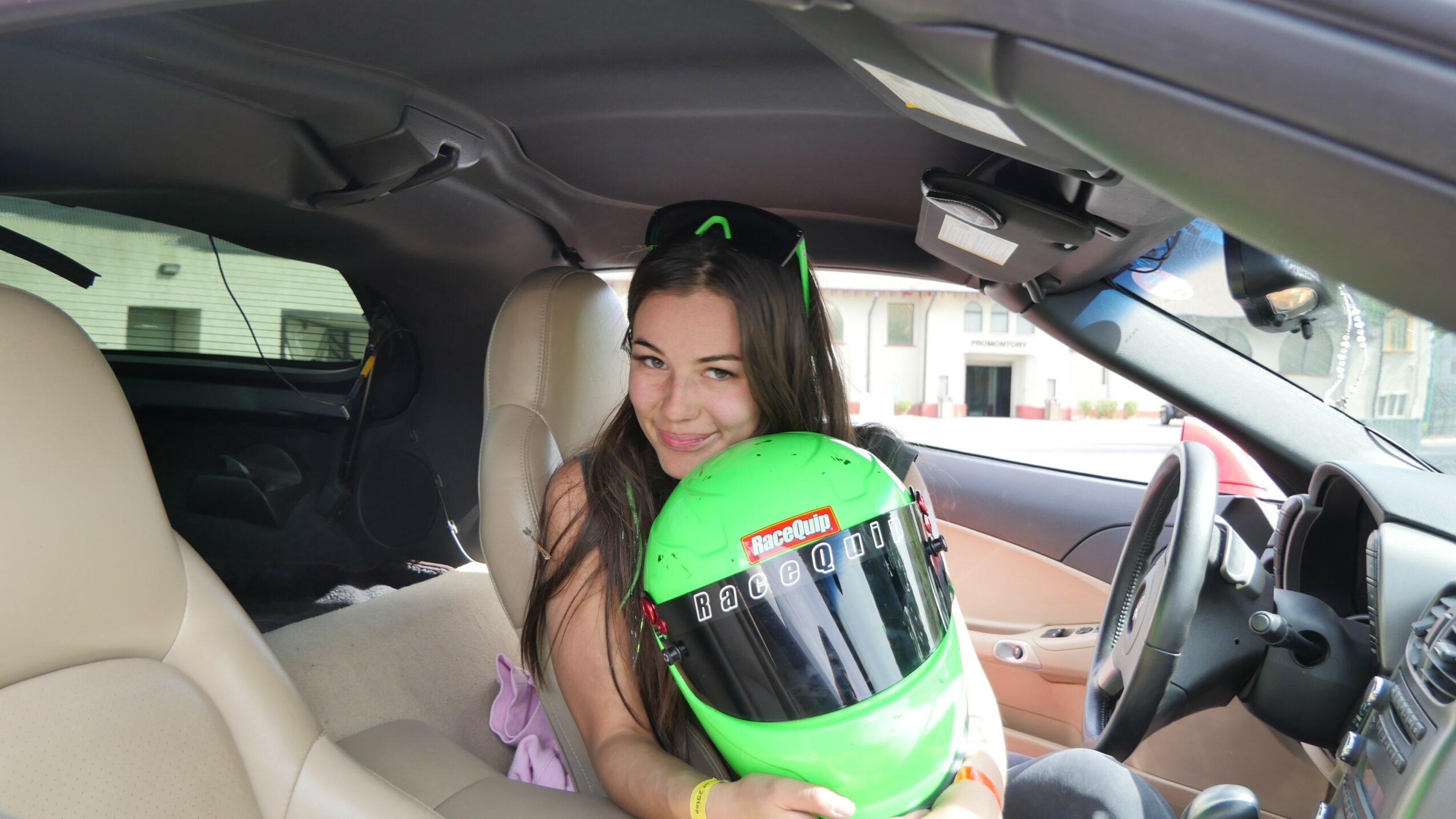 Young woman sitting in car holding a bright green motorcycle helmet, smiling, with beige car interior visible.