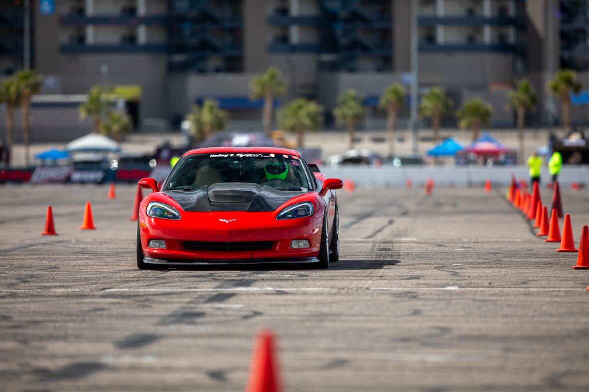 Red sports car driving through an obstacle course marked by orange cones, with buildings and people in the background.