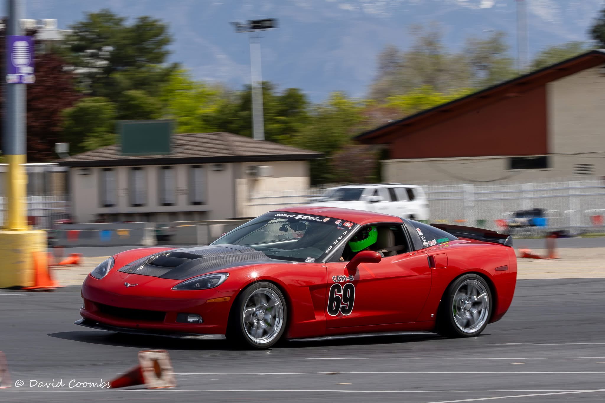 Red race car with number 69 on side driving on track, background buildings and trees, sunny day