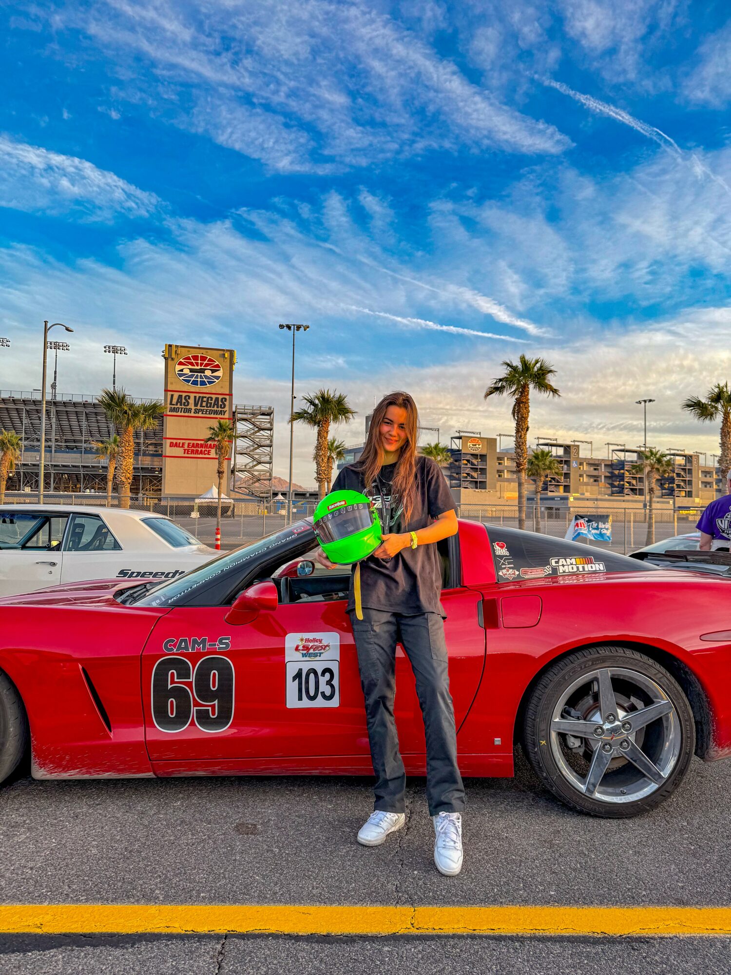 Woman standing next to a red race car holding a green helmet, in a parking lot with palm trees and blue sky.