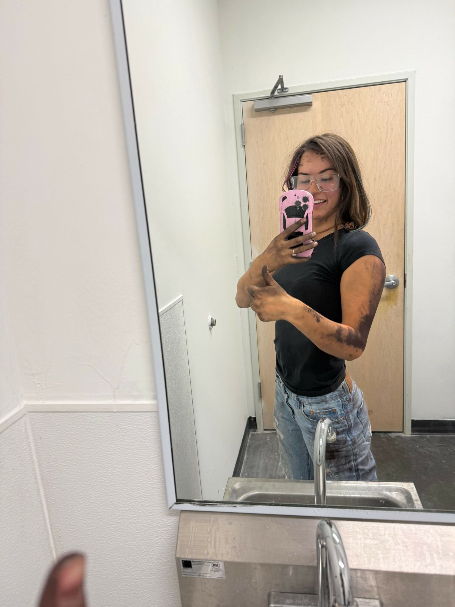 Woman taking a mirror selfie in a bathroom, holding a pink phone, with a sink and mirror visible.
