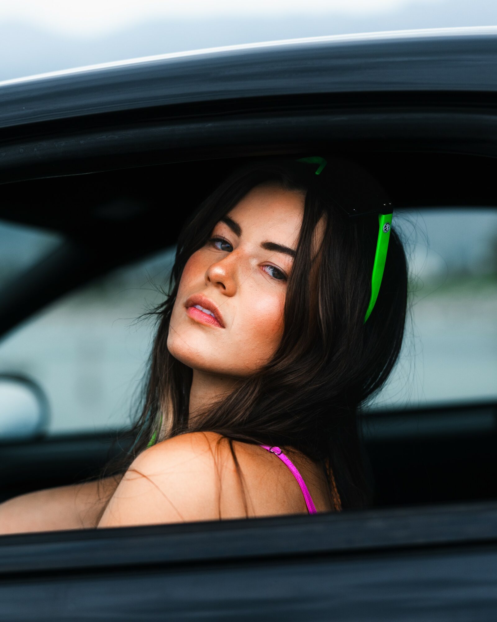 Woman with long dark hair wearing headphones, looking out of car window, with a neutral expression.