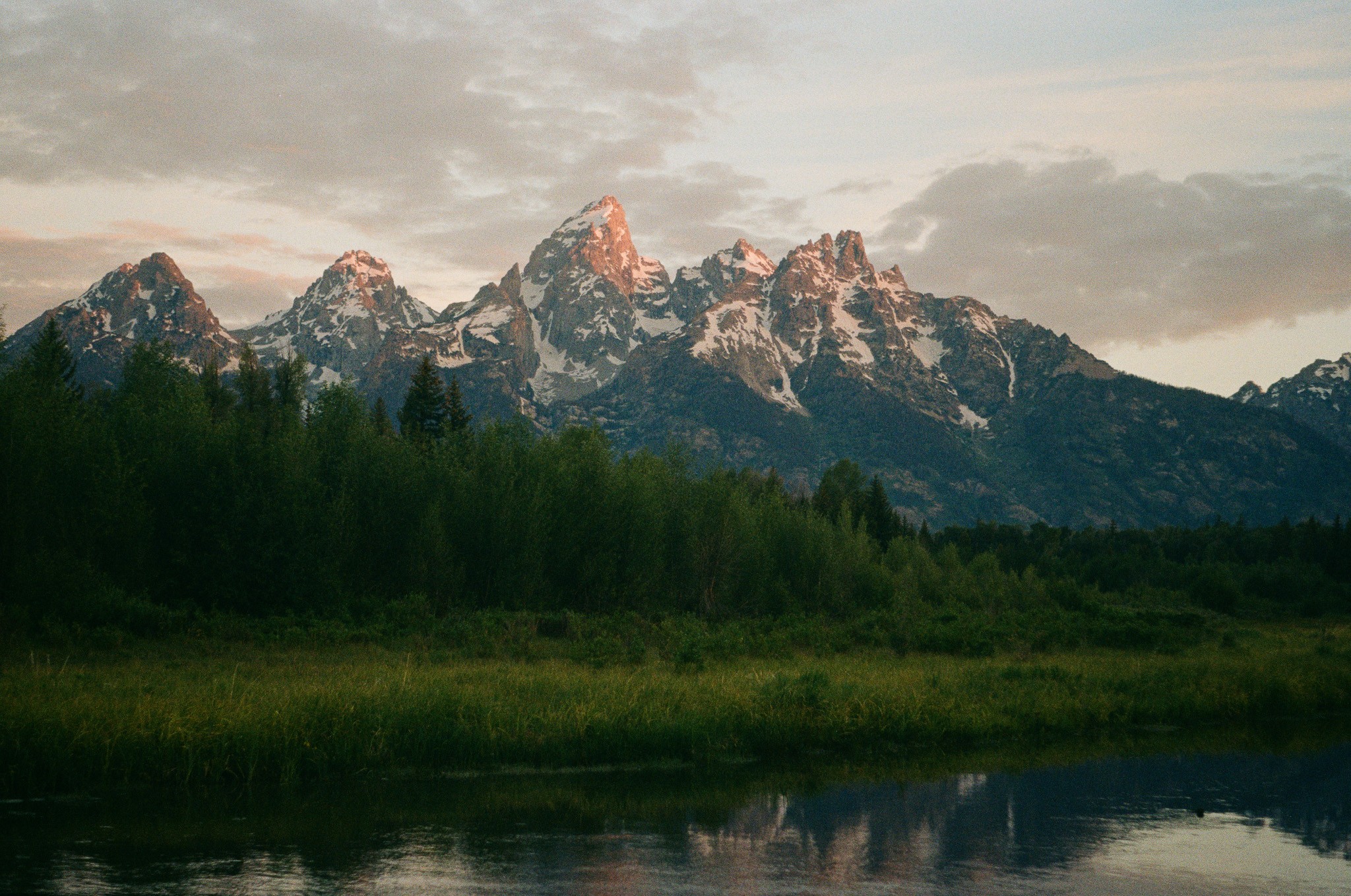 Mountain range with snow-capped peaks, forested foreground, and a body of water in the lower part of the image.