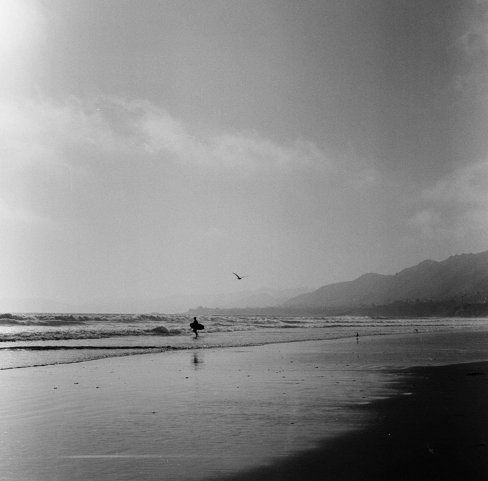 Person walking along the beach with waves and mountains in the background, cloudy sky, black and white image.