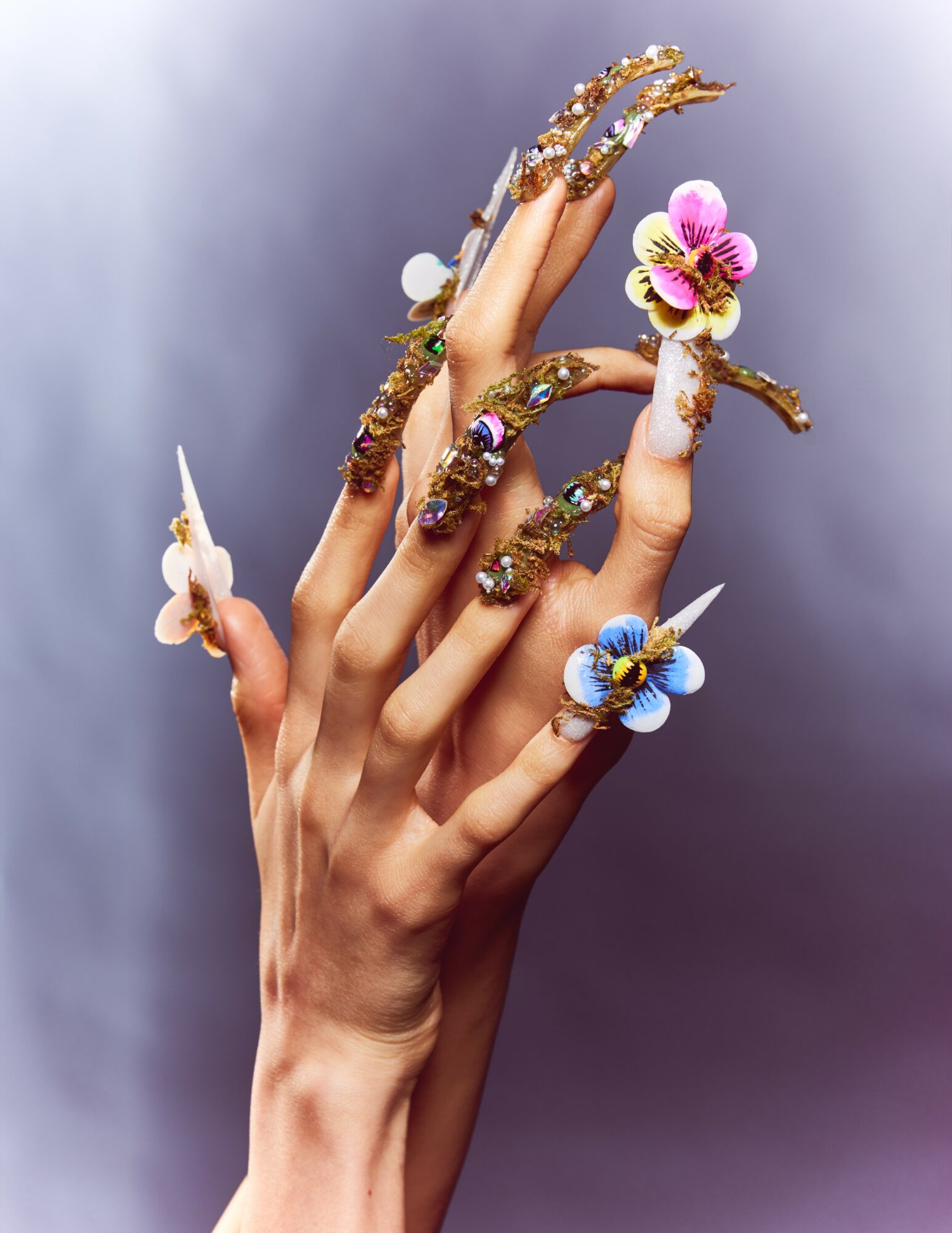 Hand with decorated nails and colorful flowers on fingers, against a blurred background.