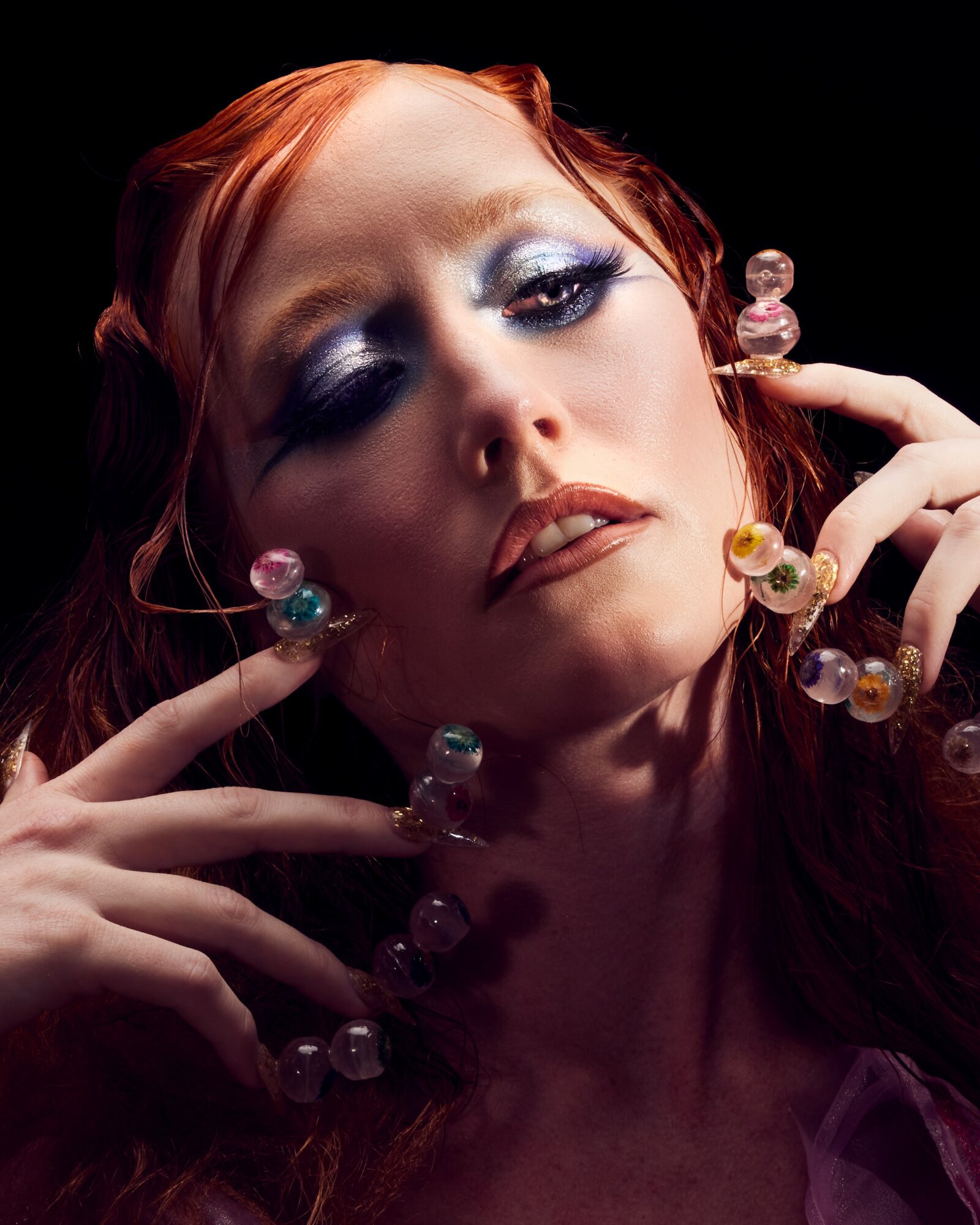 Close-up of a woman with red hair and dramatic makeup, holding colorful beads near her face.