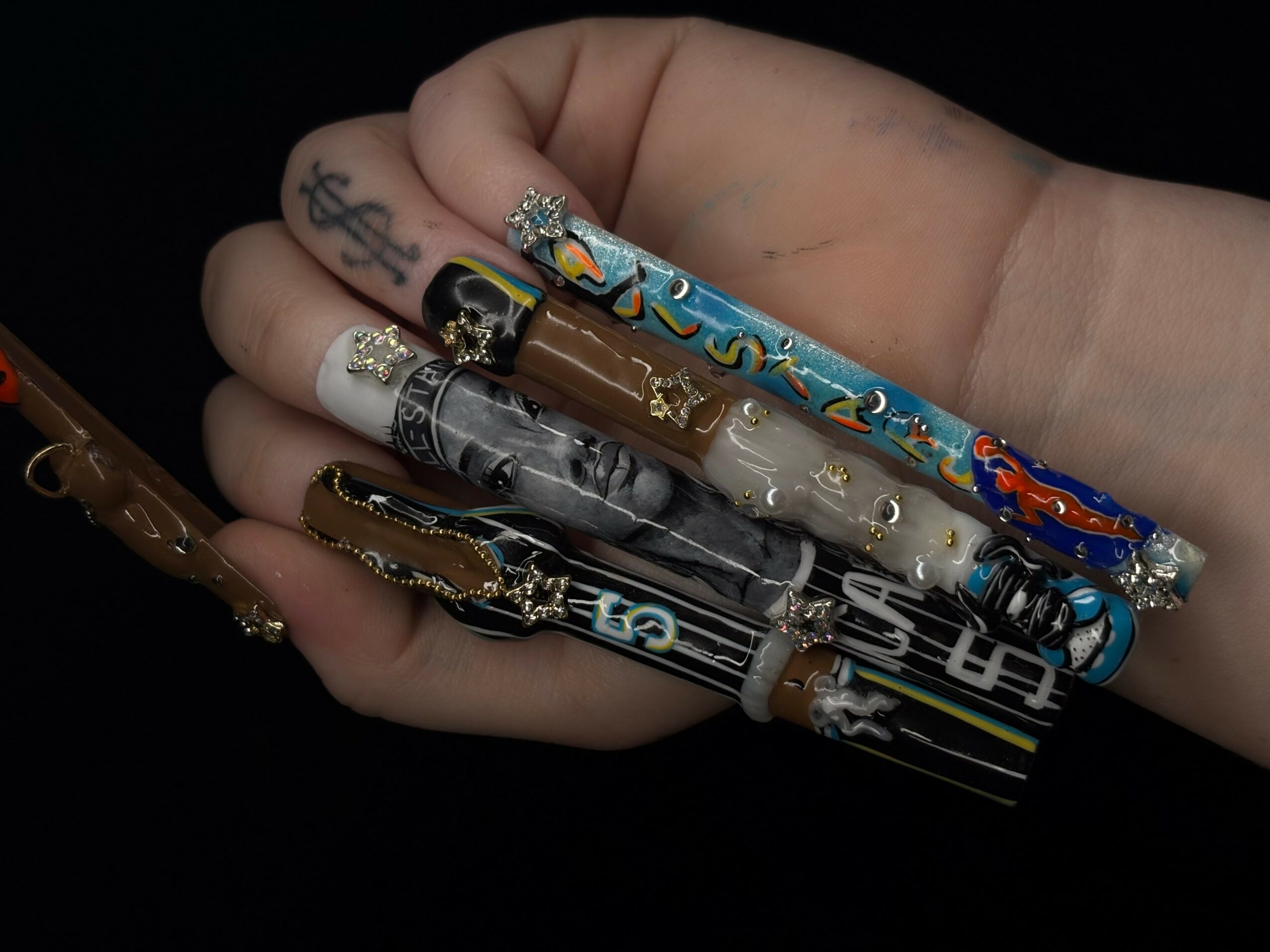 Hand holding five decorated pens with various designs and colors, against a black background.