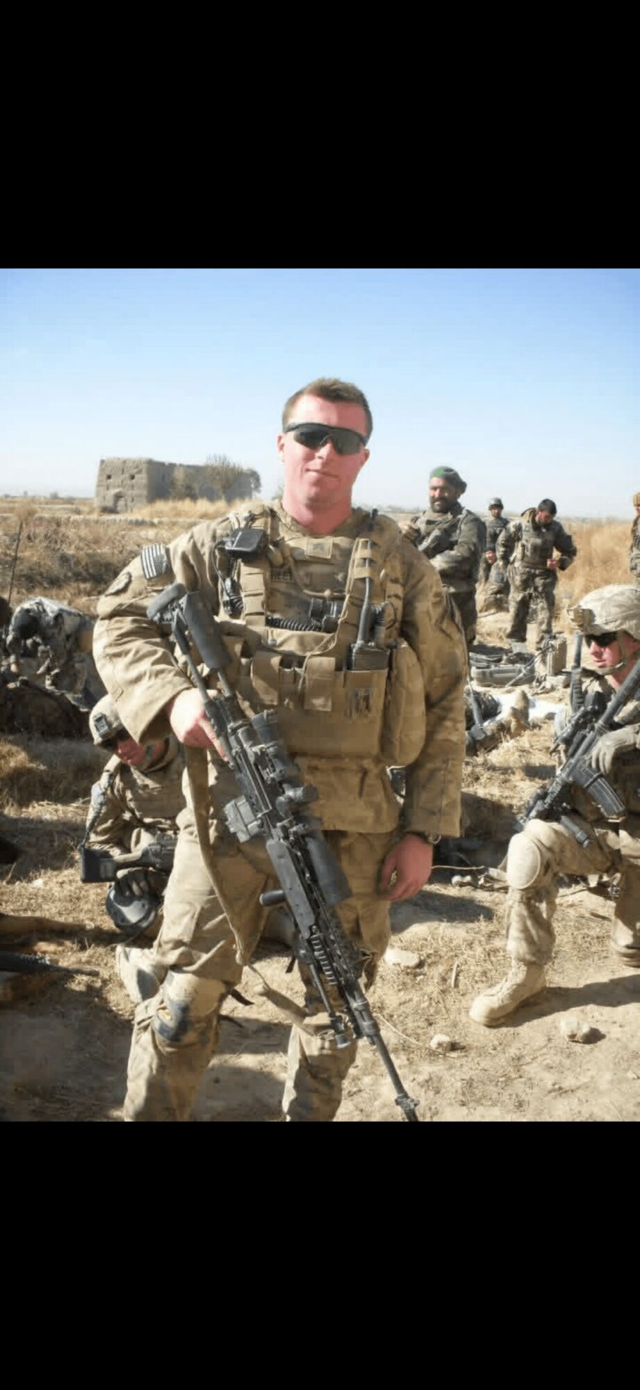 Soldier in desert uniform holding a rifle, with other soldiers in background, outdoors under clear sky.