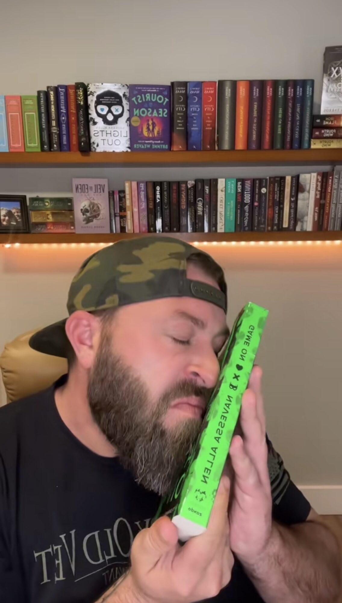 Man with a beard and camouflage cap holding a green book with closed eyes, in a room with bookshelves and warm lighting