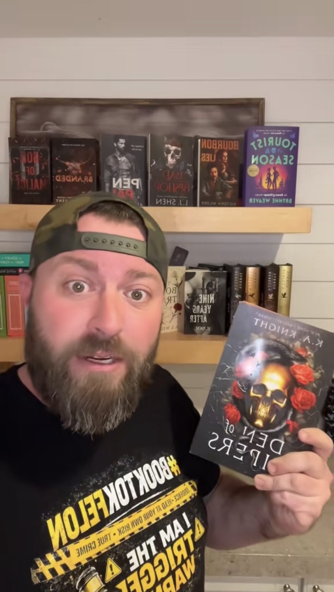 Man with a beard and cap holding a book titled 'Death of the Flesh' in front of bookshelves.