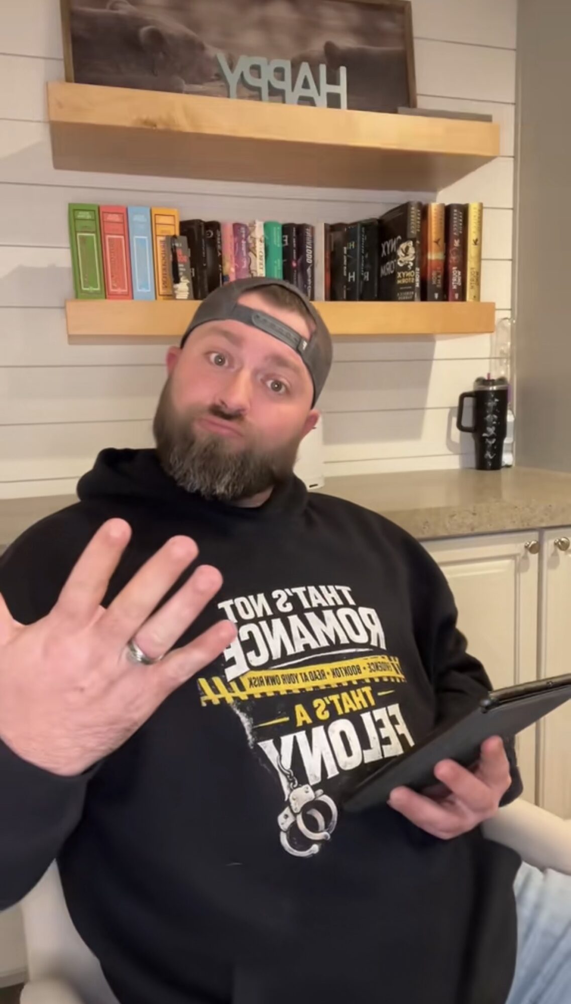 Bearded man wearing a black hoodie and cap, holding a tablet, waving, in a room with bookshelves and a countertop.